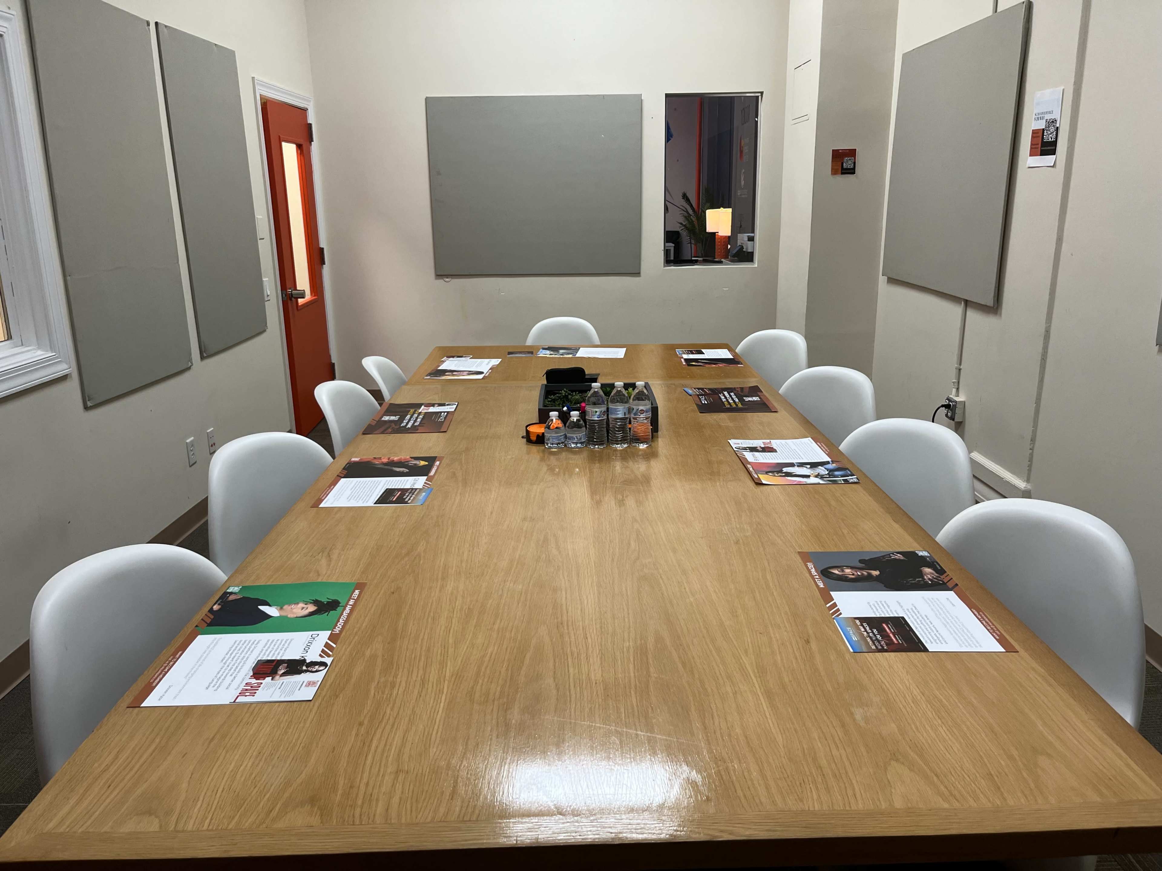 A long wooden table is set with paper materials and water bottles in a brightly lit meeting room with white walls and a red door.