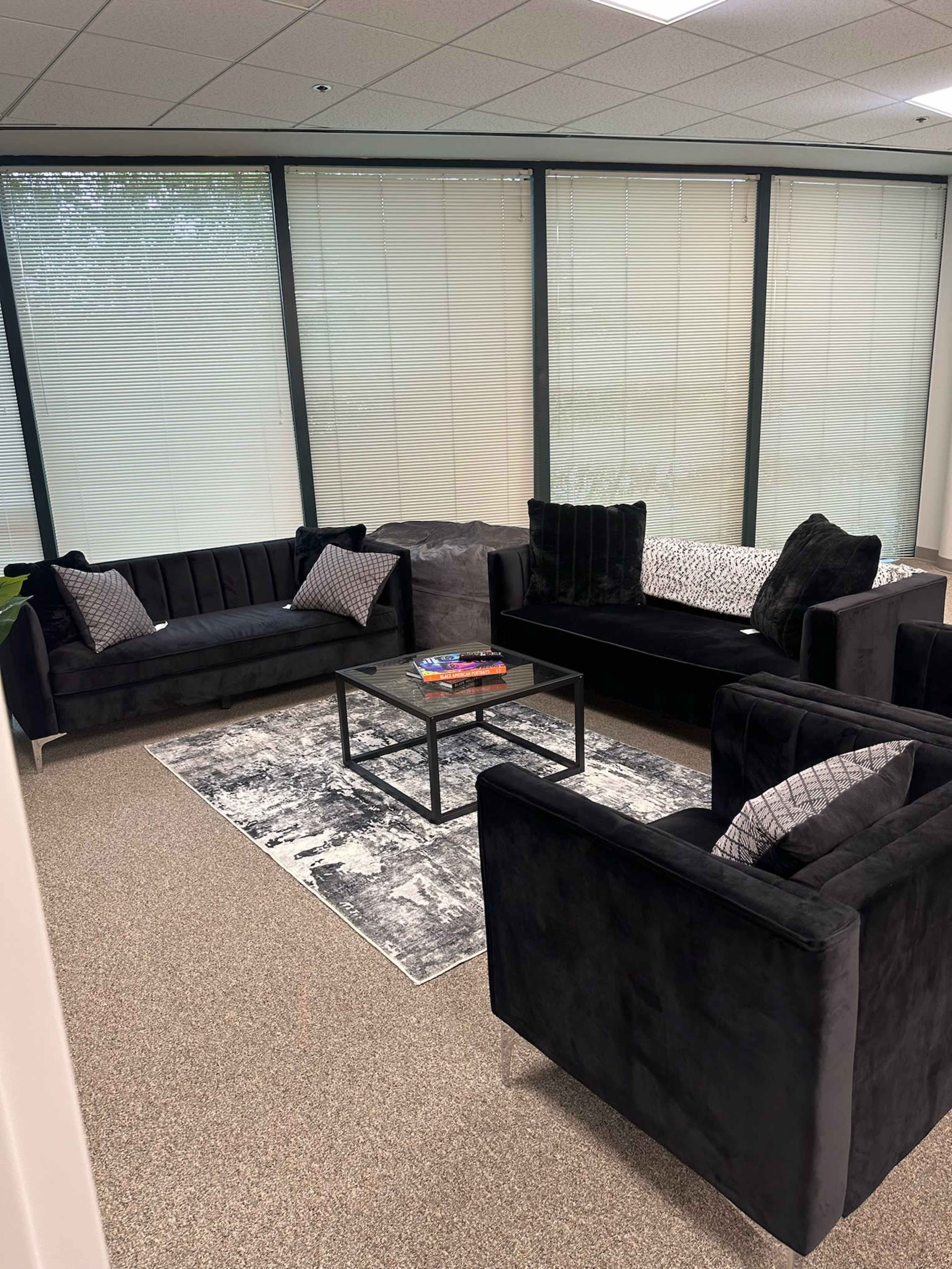 The image shows a modern office lounge area with black upholstered couches and armchairs arranged around a glass coffee table on a patterned rug.