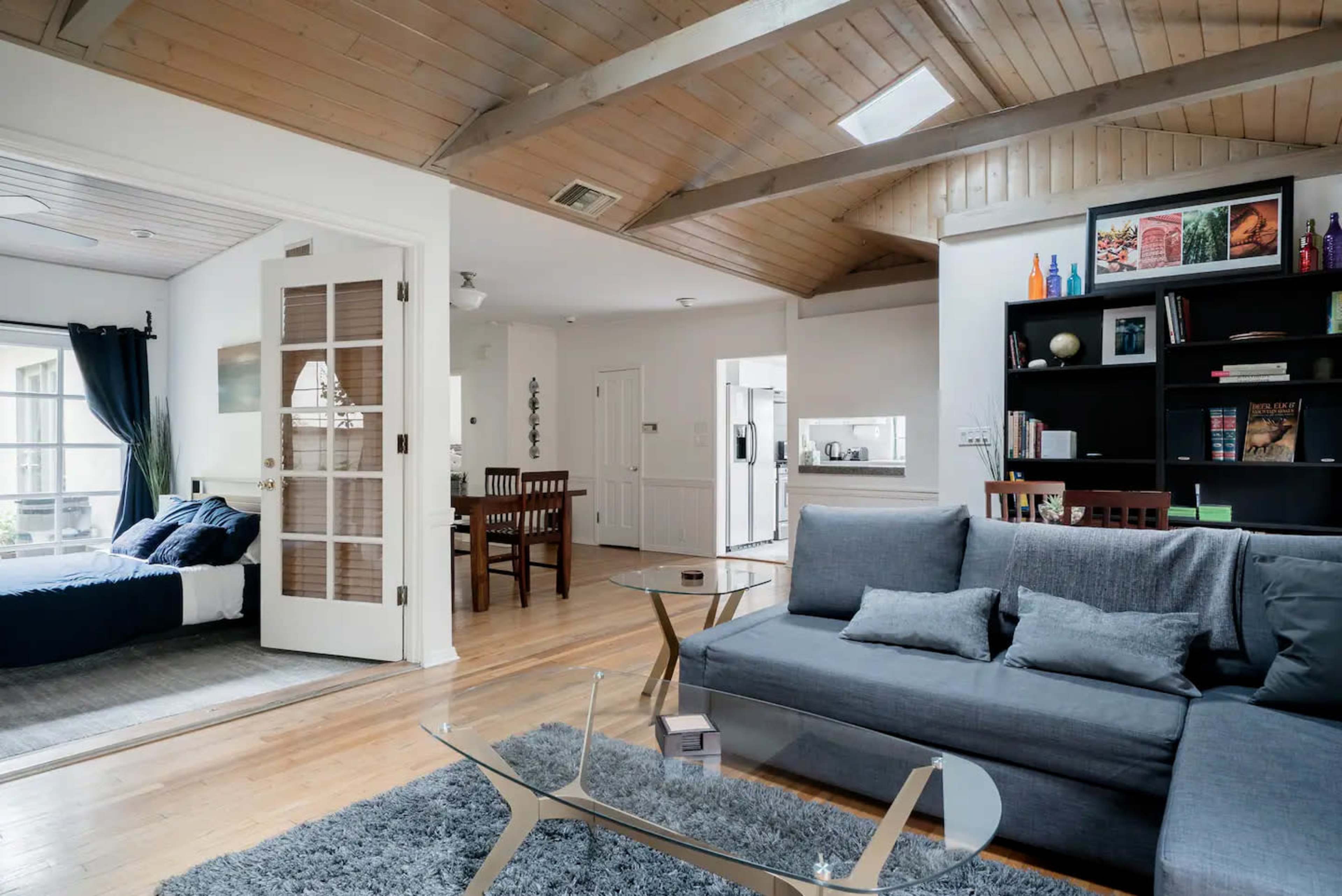 The image shows a modern living space featuring a gray sofa, a glass coffee table, wooden floors, and an adjacent dining area with a wooden table and chairs.
