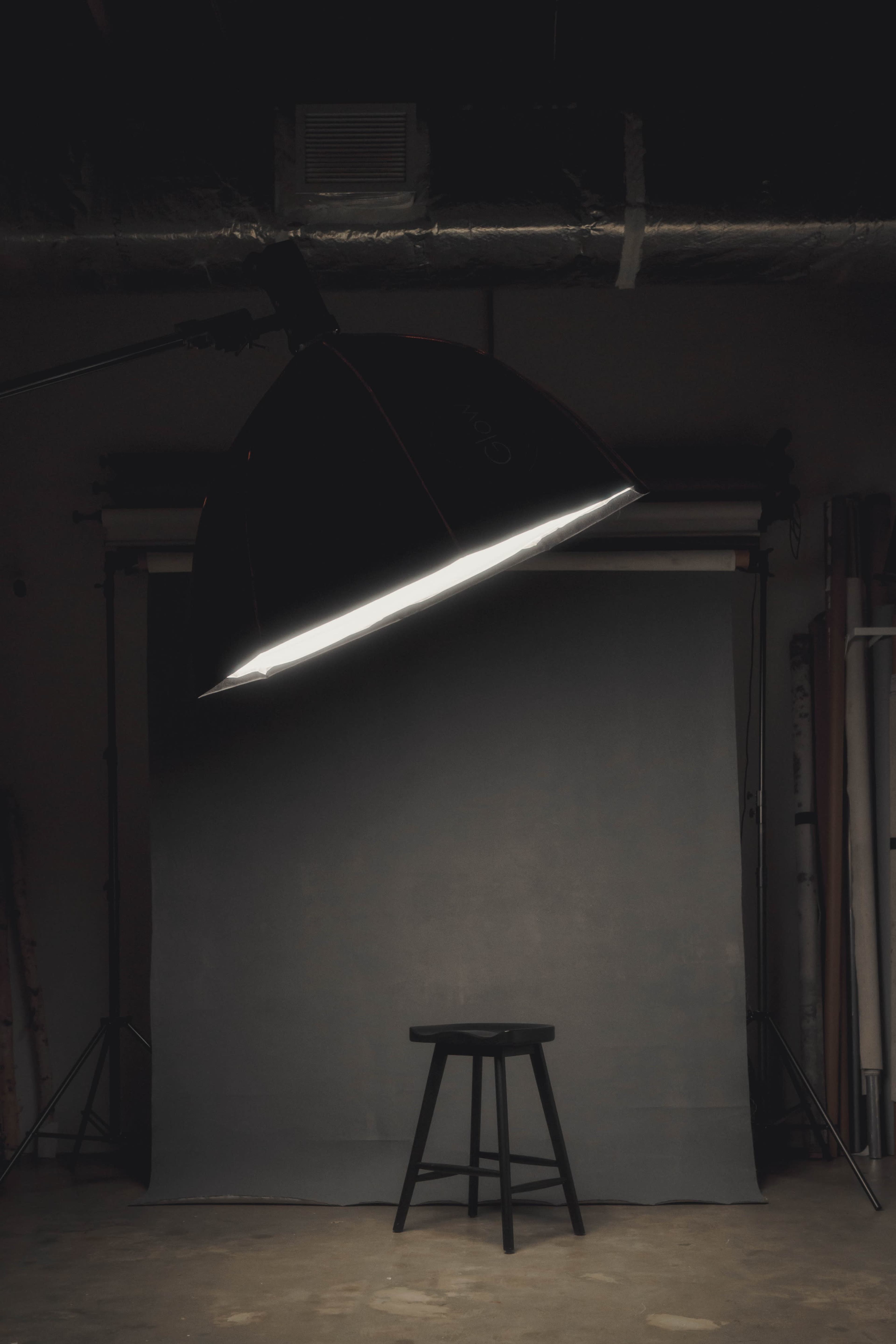 A large softbox light is positioned above a black stool in front of a gray backdrop in a photography studio.