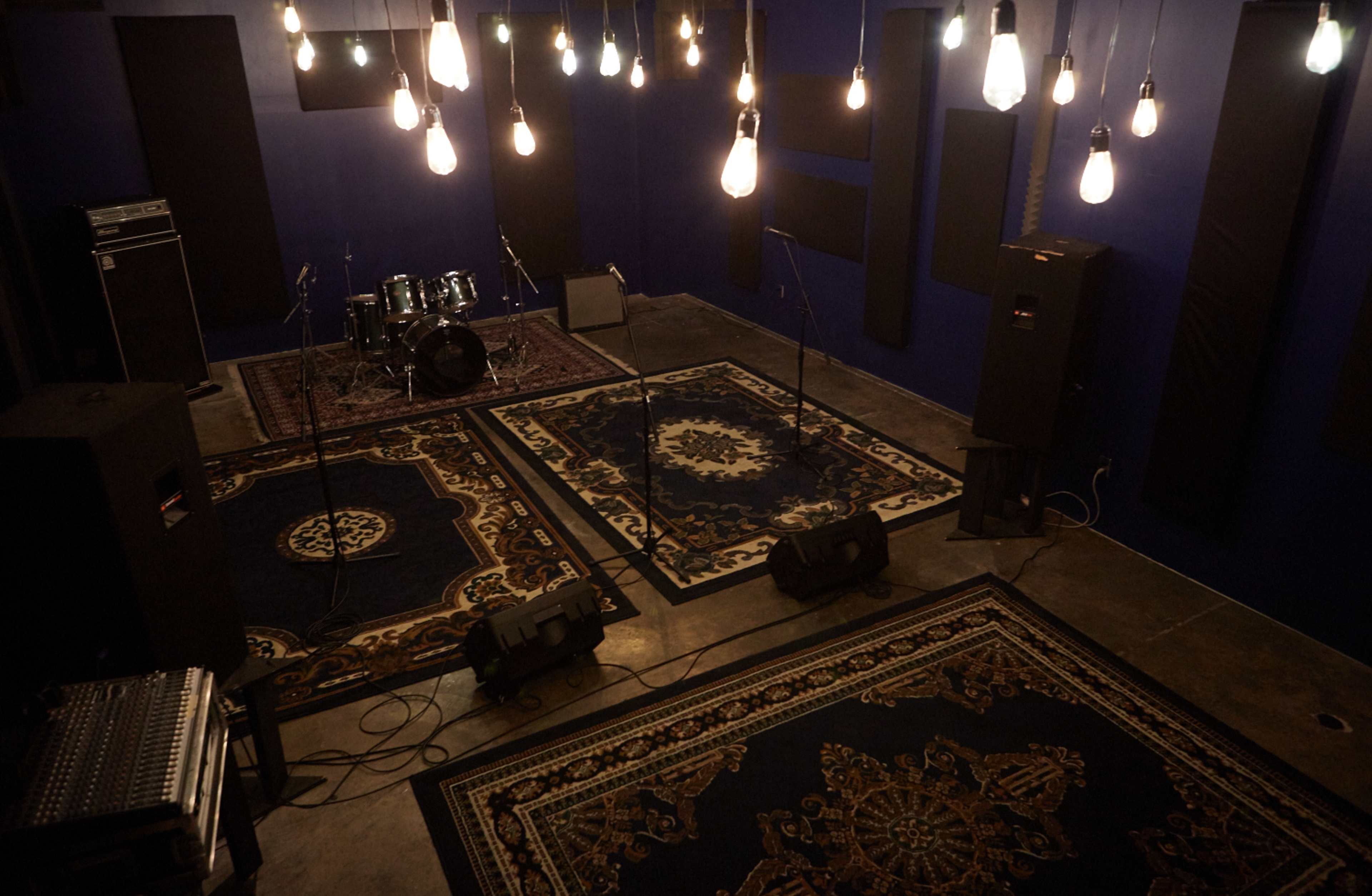 A music rehearsal space features a drum set, microphones on stands, and illuminated by hanging light bulbs, with rugs on a concrete floor.