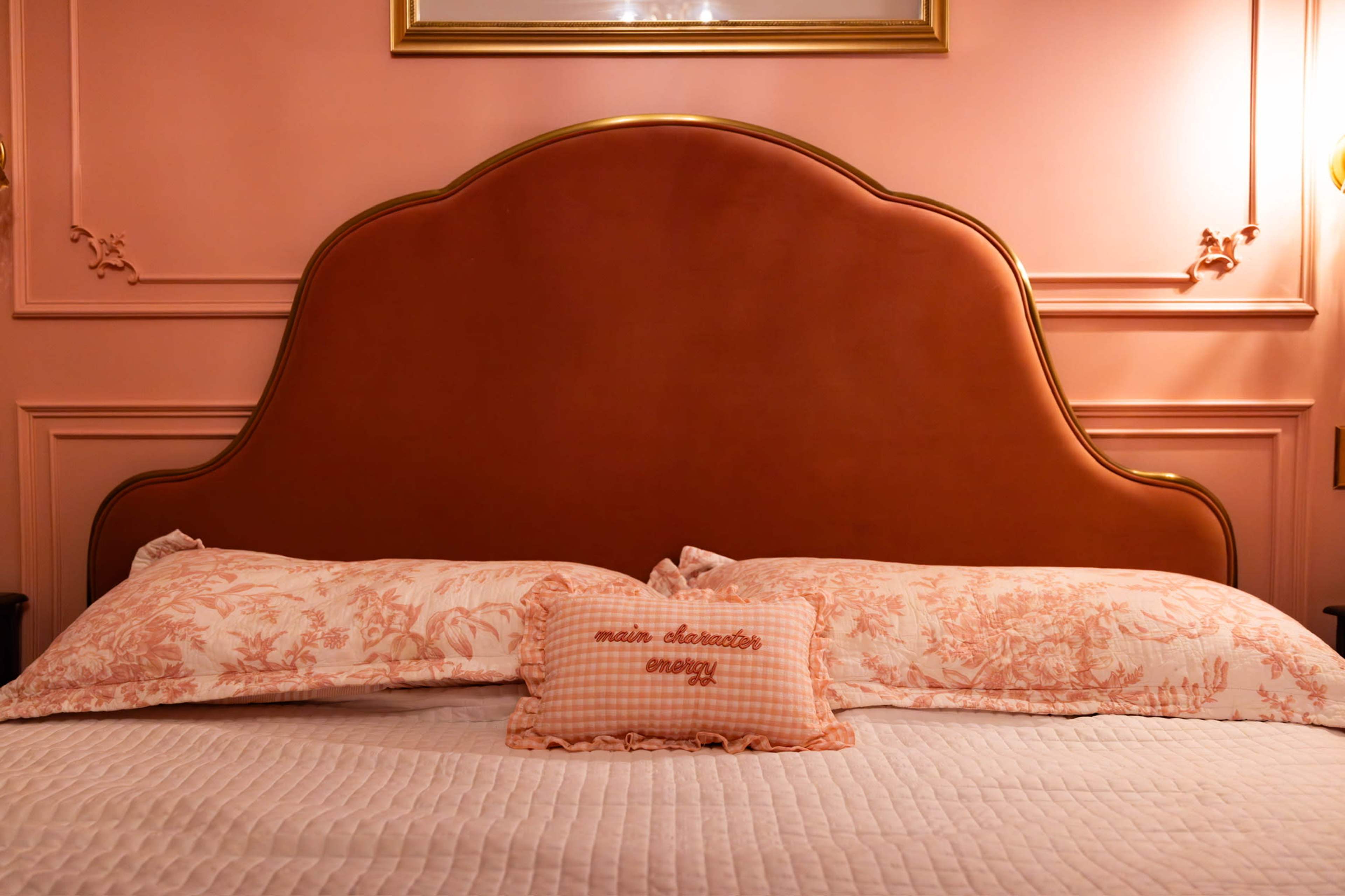 The image shows a bed with a brown leather headboard, two decorative pillows, and a smaller embroidered pillow in front, against a pink wall.