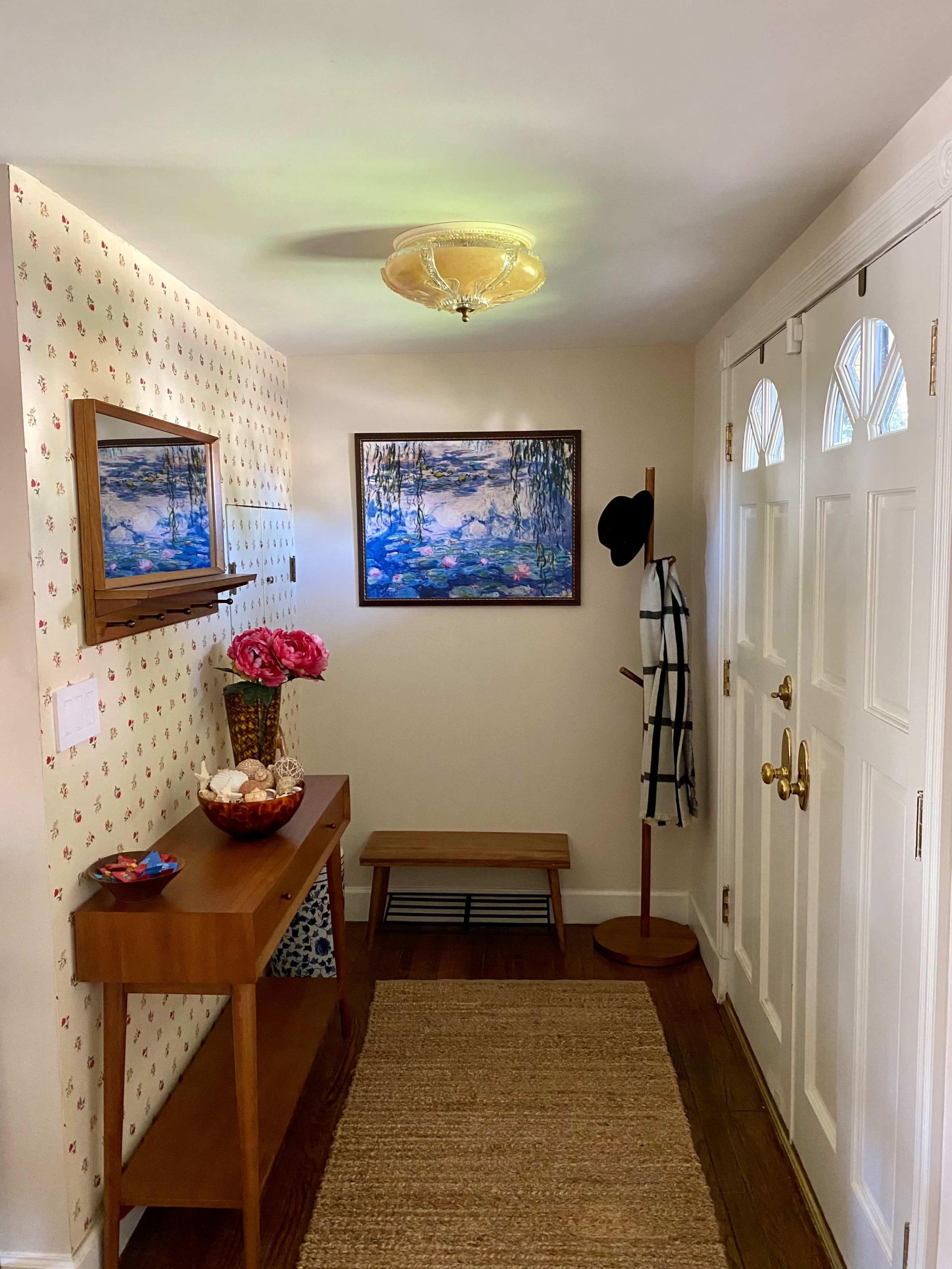 The image shows a well-lit entryway with a wooden console table, a floral arrangement, a painting on the wall, and two doors leading outside.