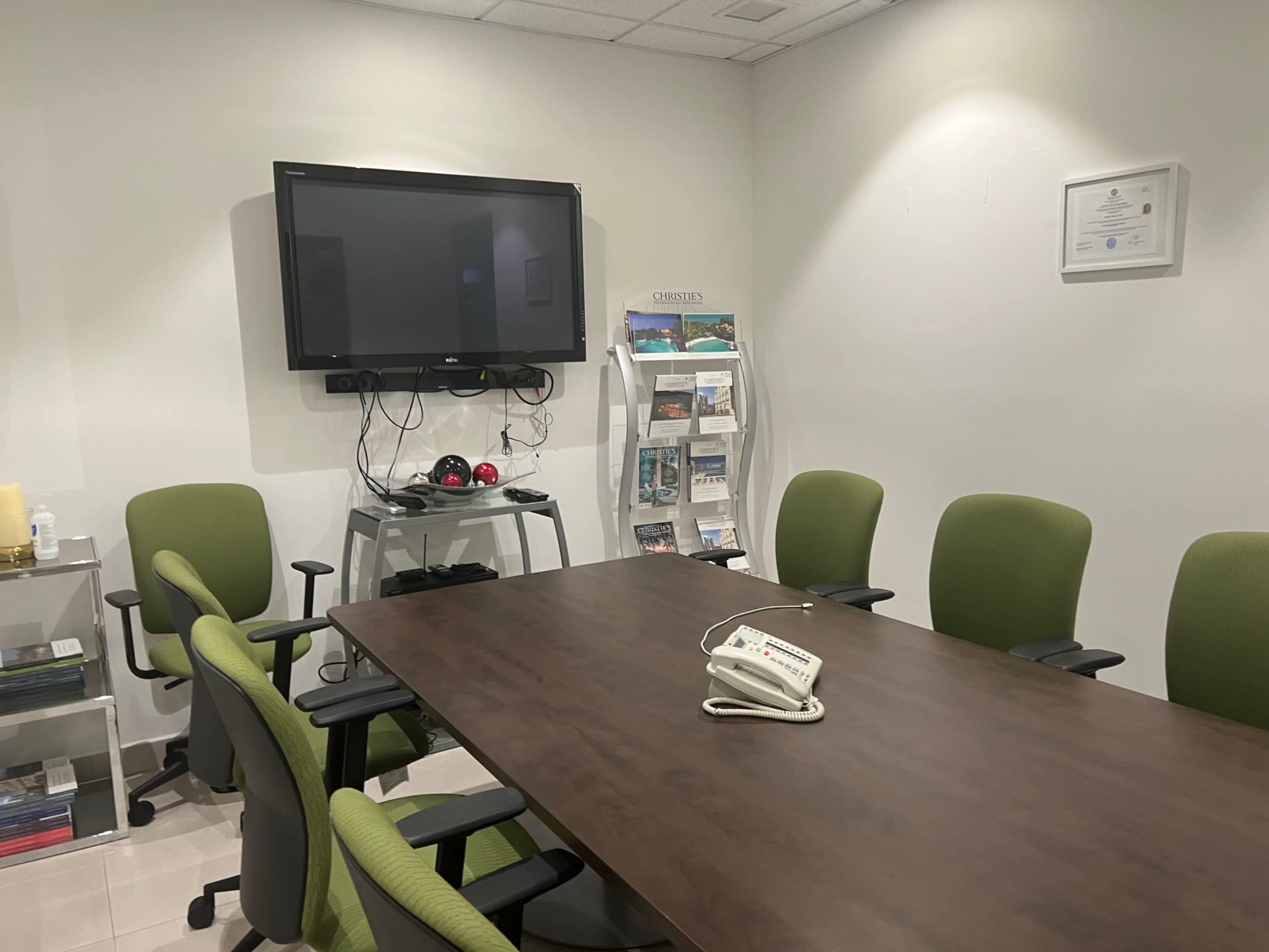 The image shows a small conference room with a wooden table, green chairs, a wall-mounted TV, and a phone on the table.