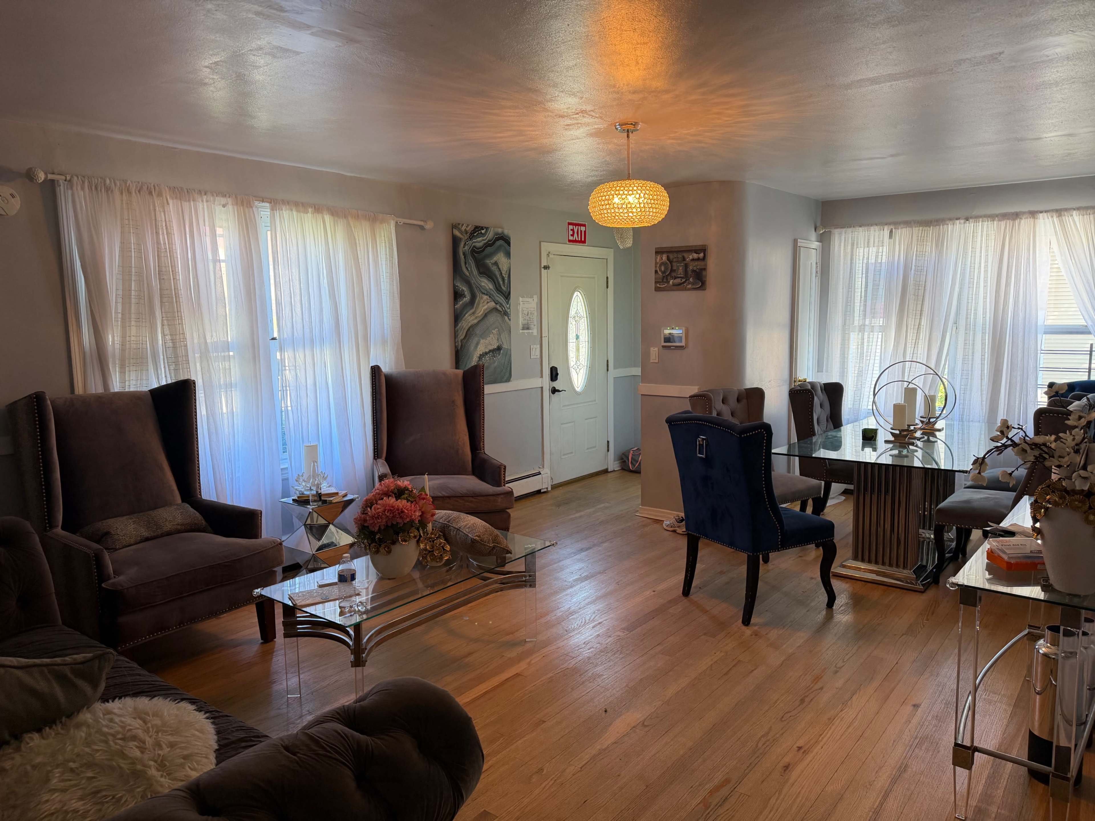 The image shows a spacious living area with a combination of upholstered chairs, a glass coffee table, and a dining table, all illuminated by natural light streaming through the sheer curtains.