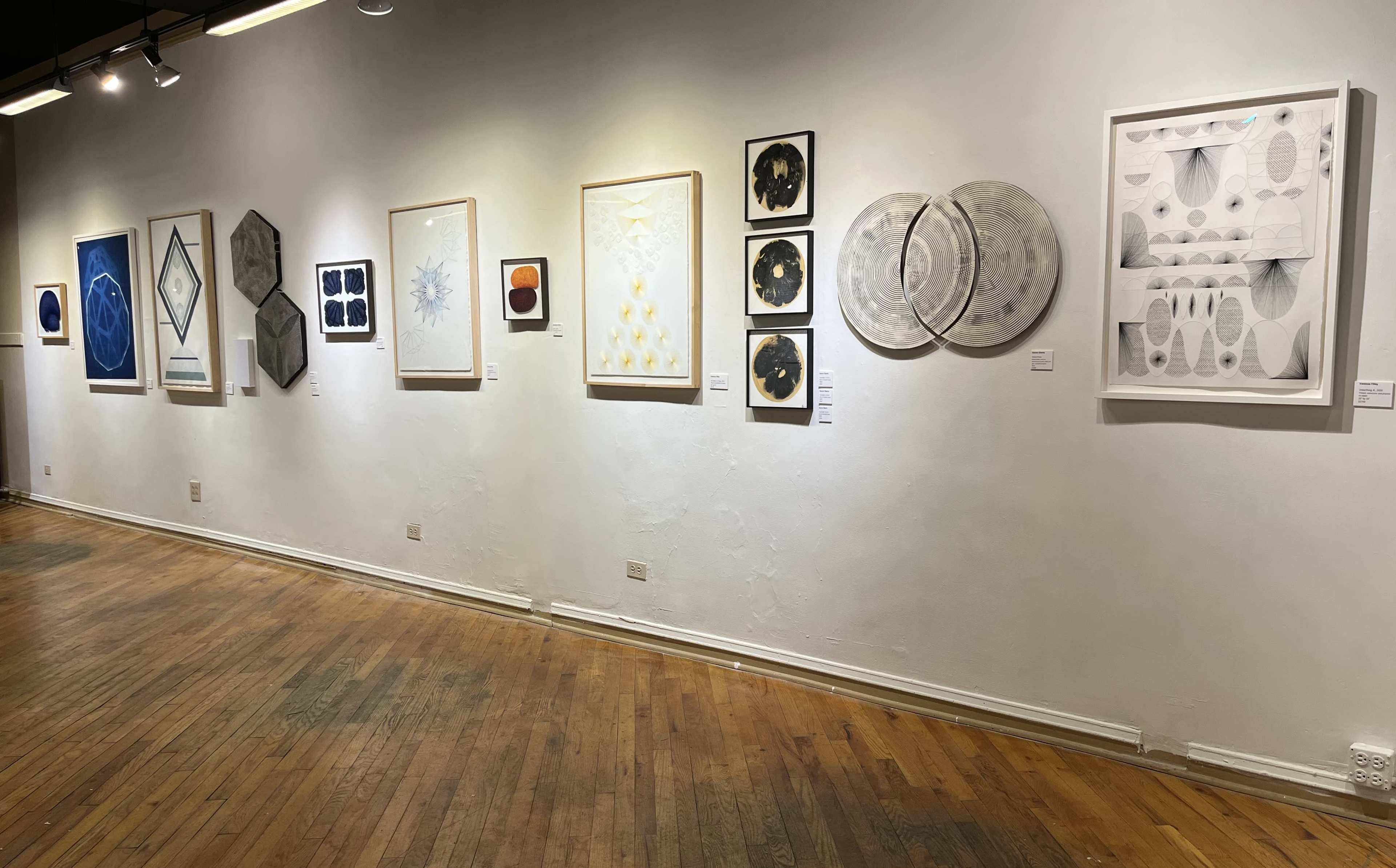 The image shows a gallery wall displaying a series of framed artworks in various styles and colors, arranged in a linear formation.