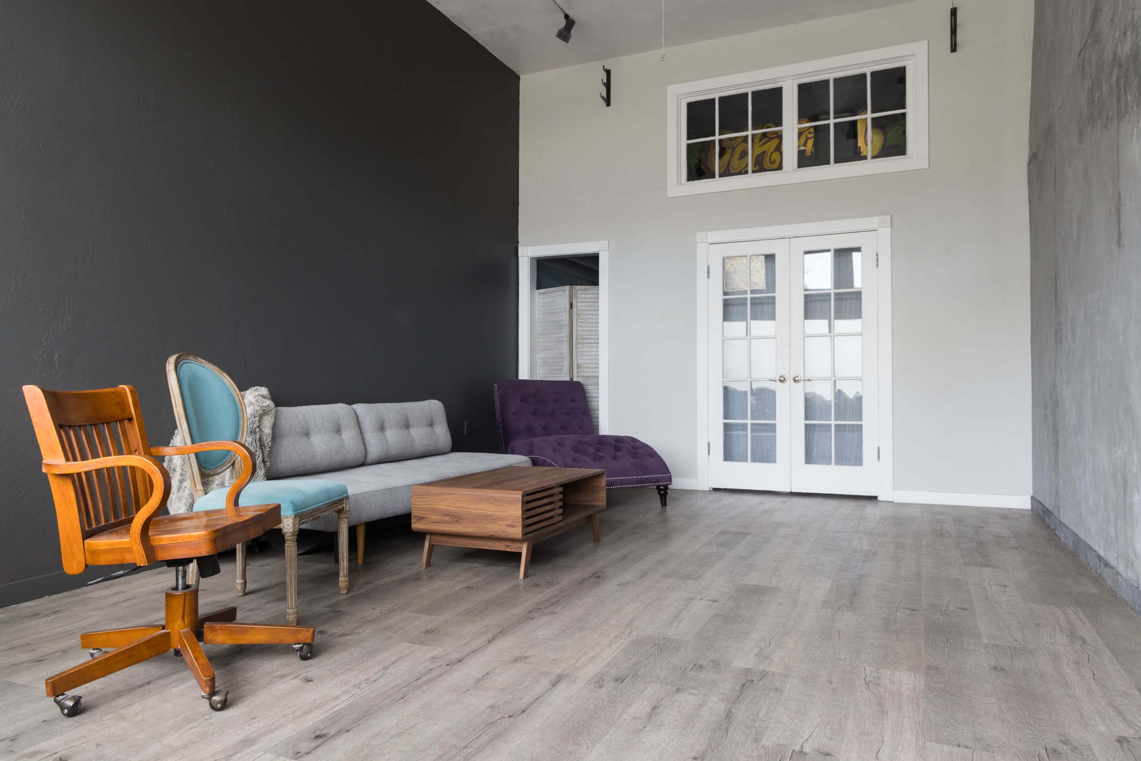 The image shows a spacious room with a black accent wall, featuring a variety of seating including a gray sofa, purple chaise lounge, wooden chair, and a coffee table, along with glass double doors leading to an outdoor area.