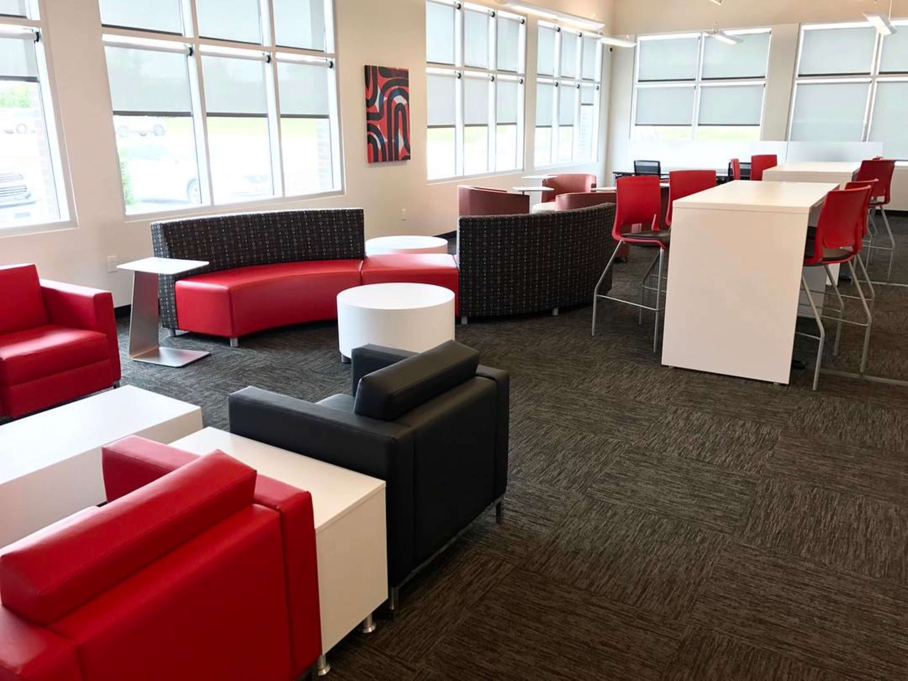 The image shows a modern lounge area with a mix of red and black seating, white tables, and large windows allowing natural light.