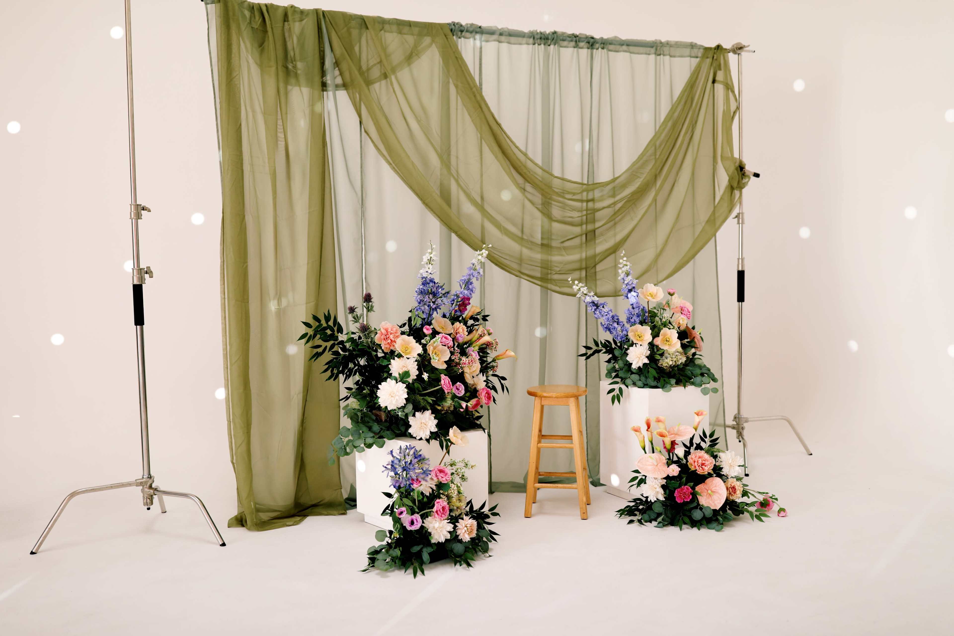 The image features a floral backdrop created with draped greenery and arrangements of flowers on two white pedestals, with a wooden stool positioned in front.