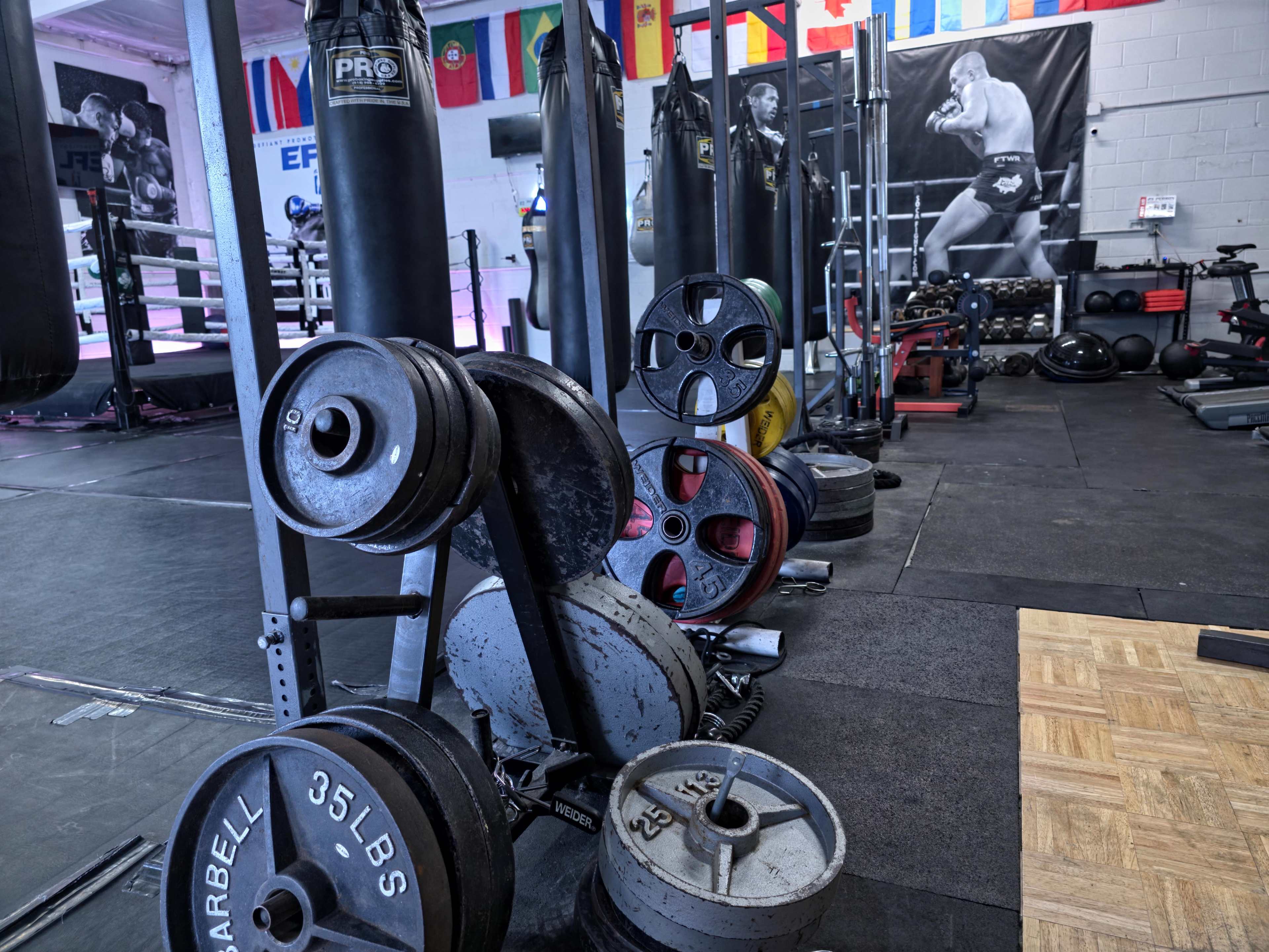 The image shows a gym interior with various weight plates stacked beside a boxing ring and exercise equipment.
