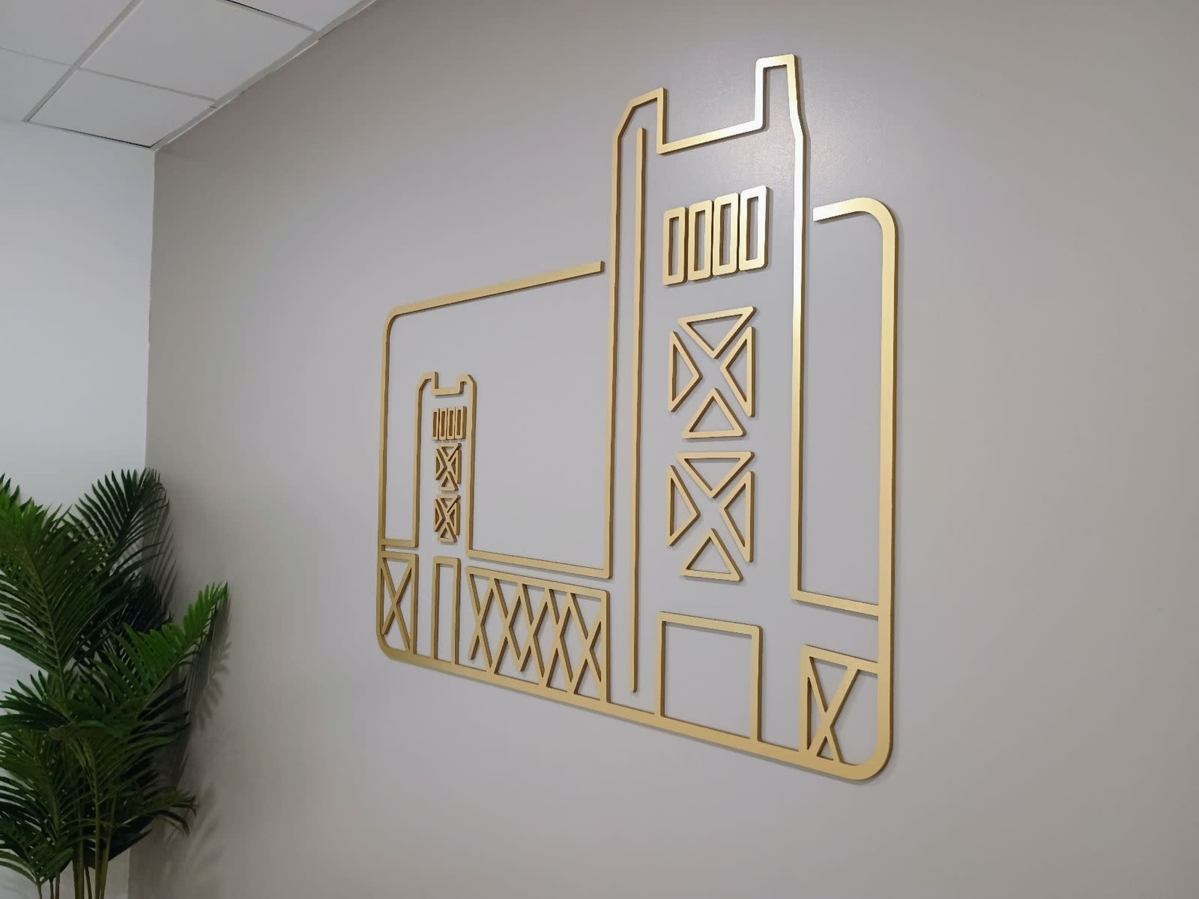 The image shows a gold metal wall decoration of a bridge design mounted on a light-colored wall, with a potted plant partially visible in the foreground.