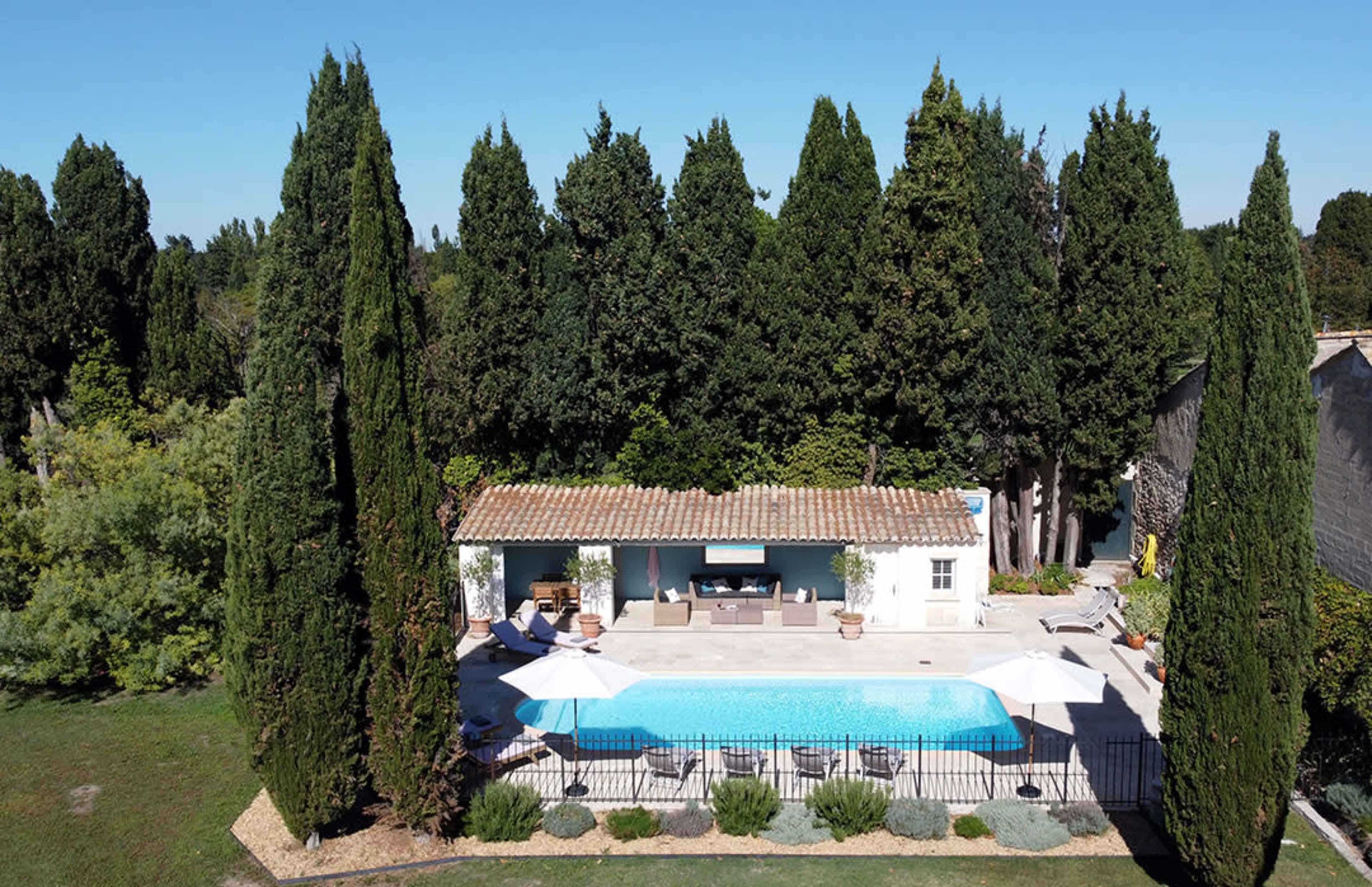A rectangular swimming pool is surrounded by tall cypress trees and a stone patio with lounge chairs and umbrellas.