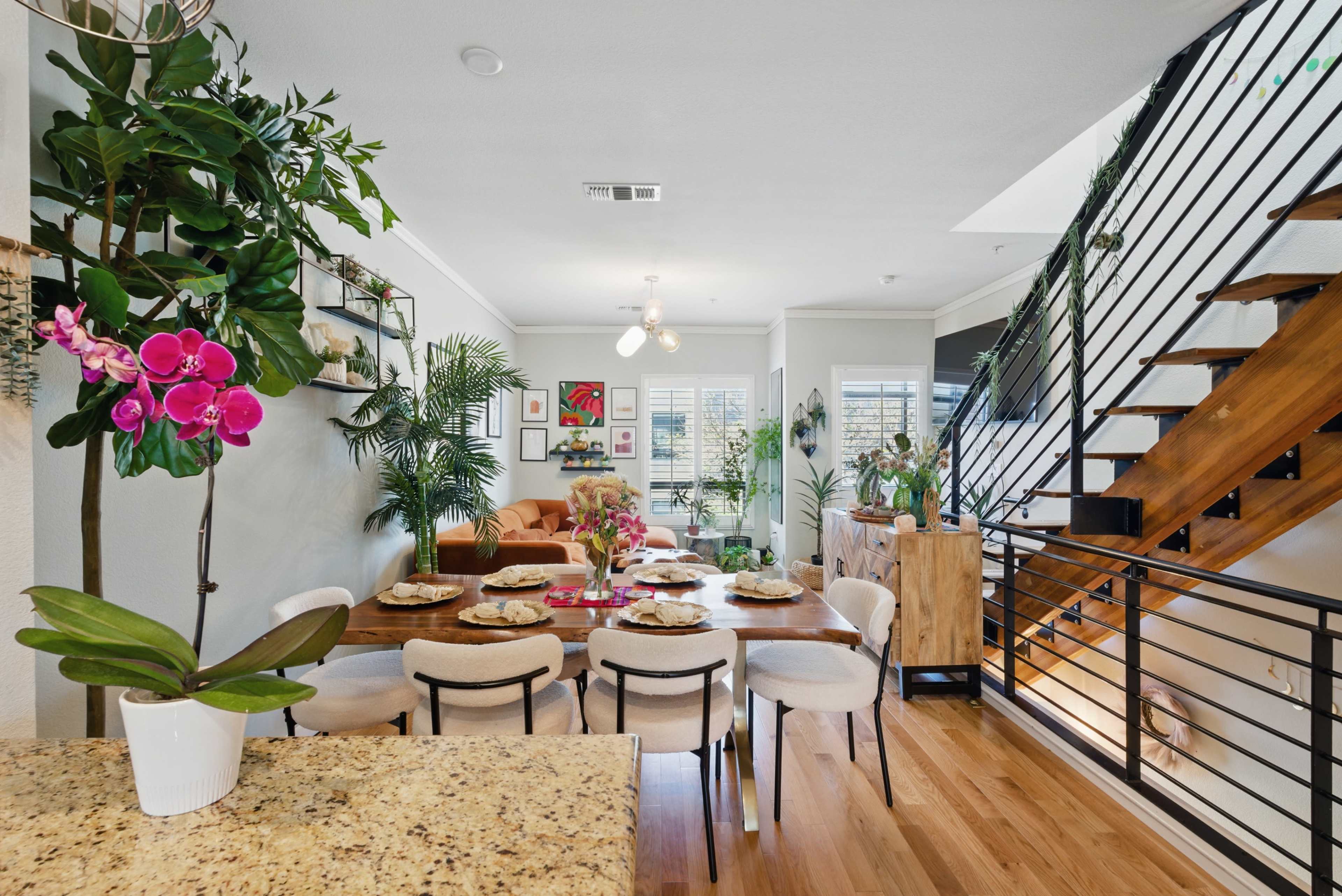 A modern, open-concept living and dining area features a wooden table set for dinner, surrounded by plants and contemporary decor, with a staircase visible in the background.