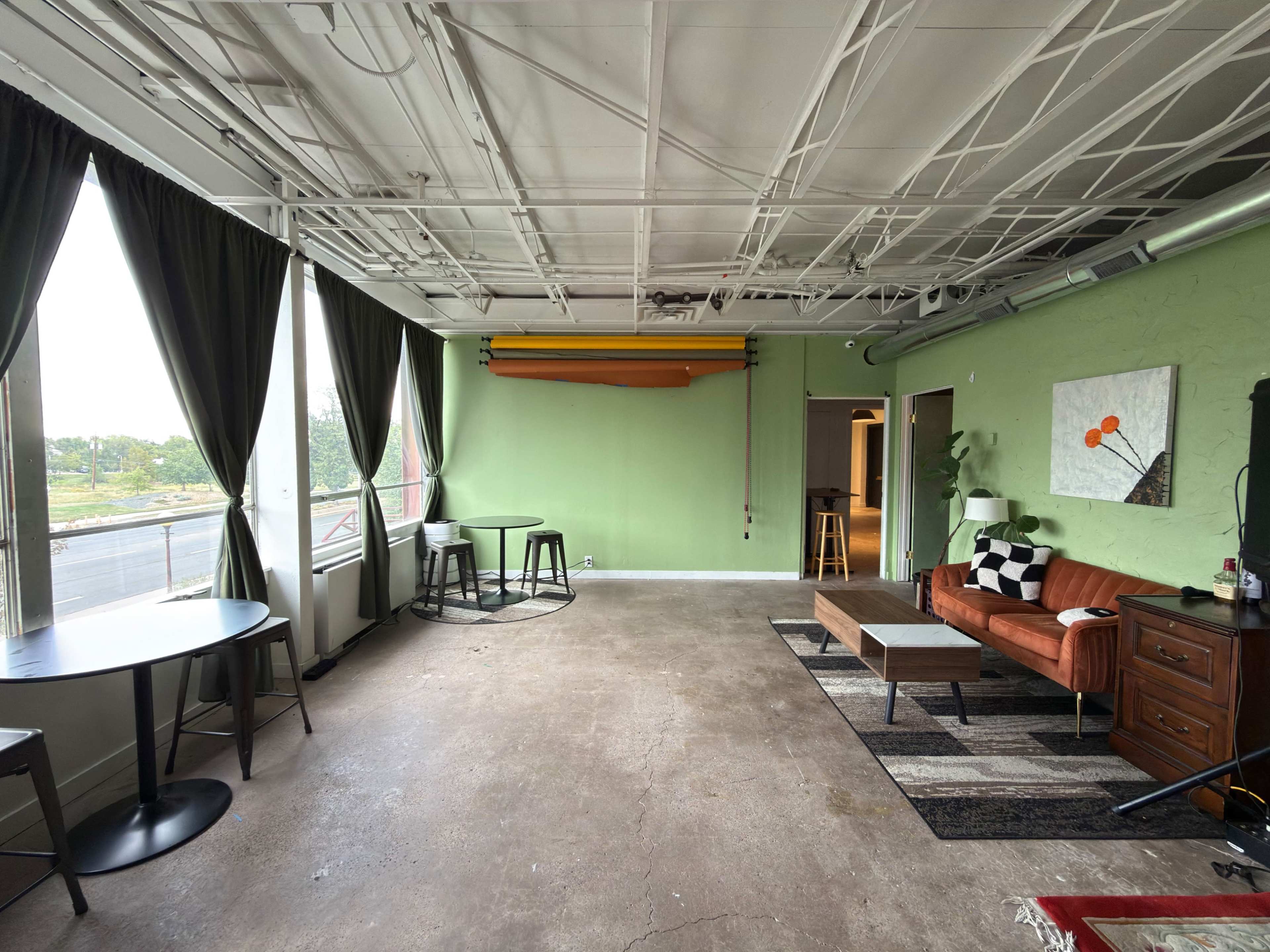 The image shows an empty room with a green wall, large windows, a brown sofa, a coffee table, and a few tables set up for seating.