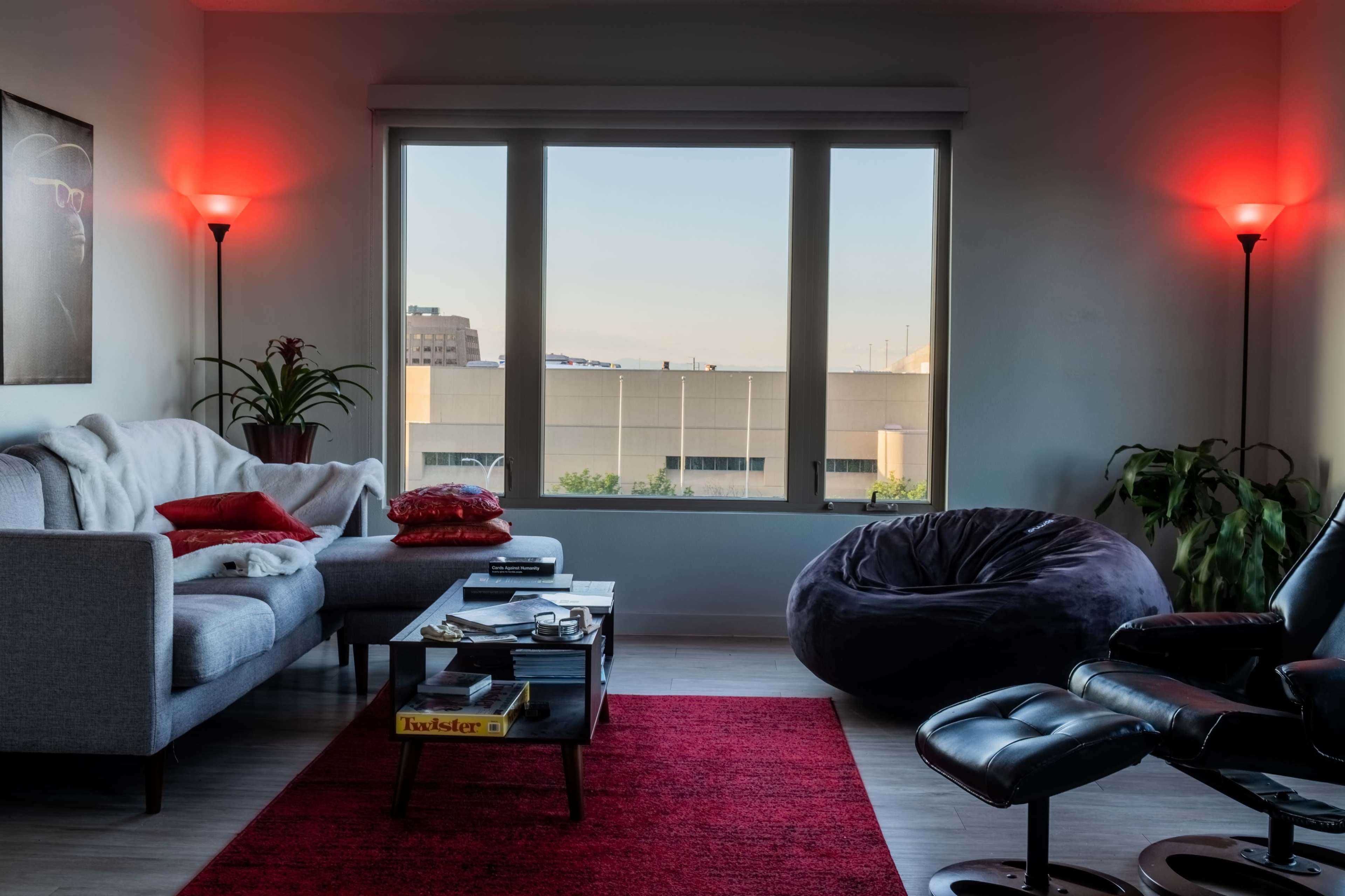 A modern living room features a gray sofa with red pillows, a glass coffee table, and a large window with red lamps illuminating the space.