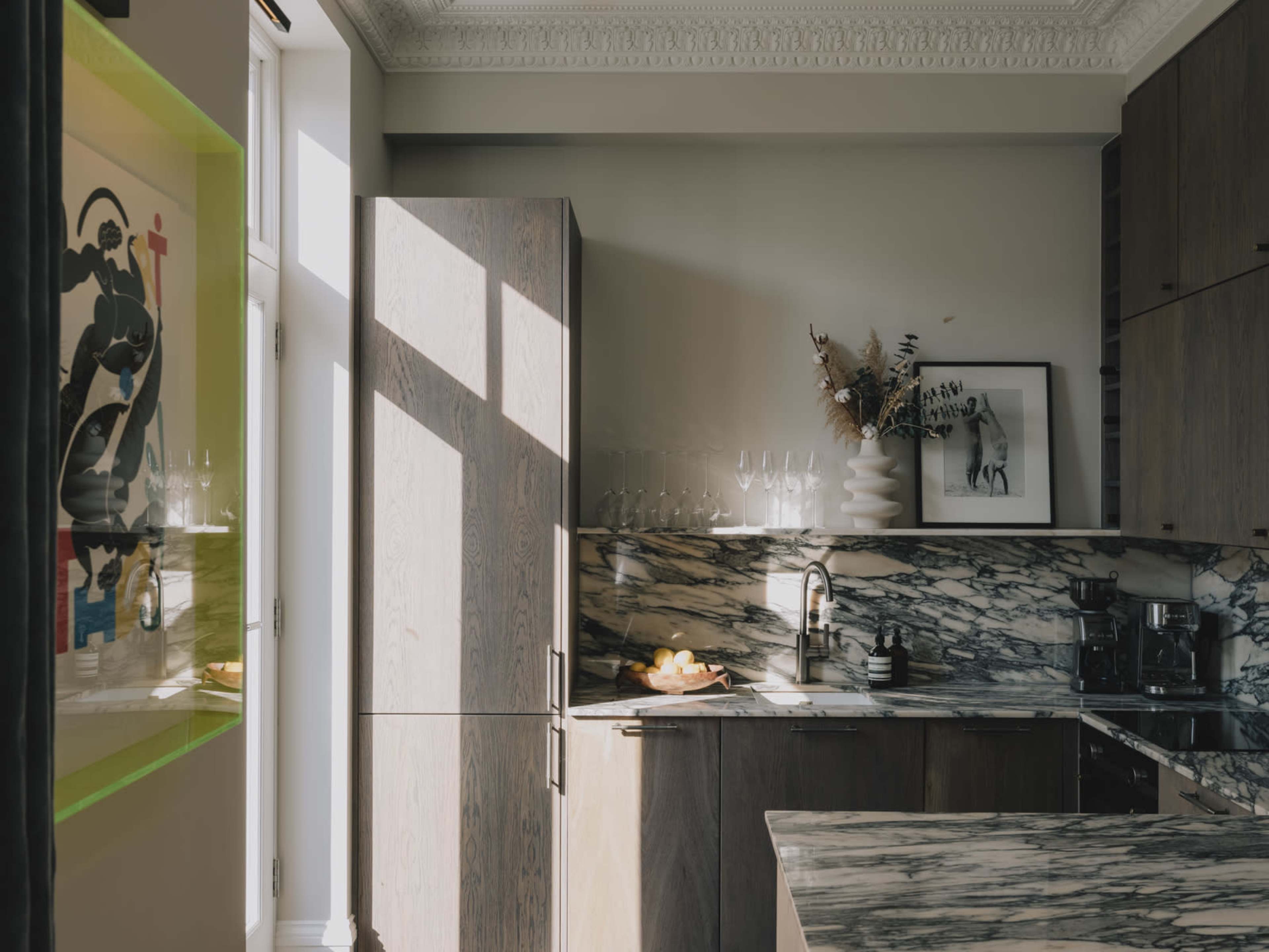 A modern kitchen features marble countertops, wooden cabinets, and large windows allowing natural light to illuminate the space.