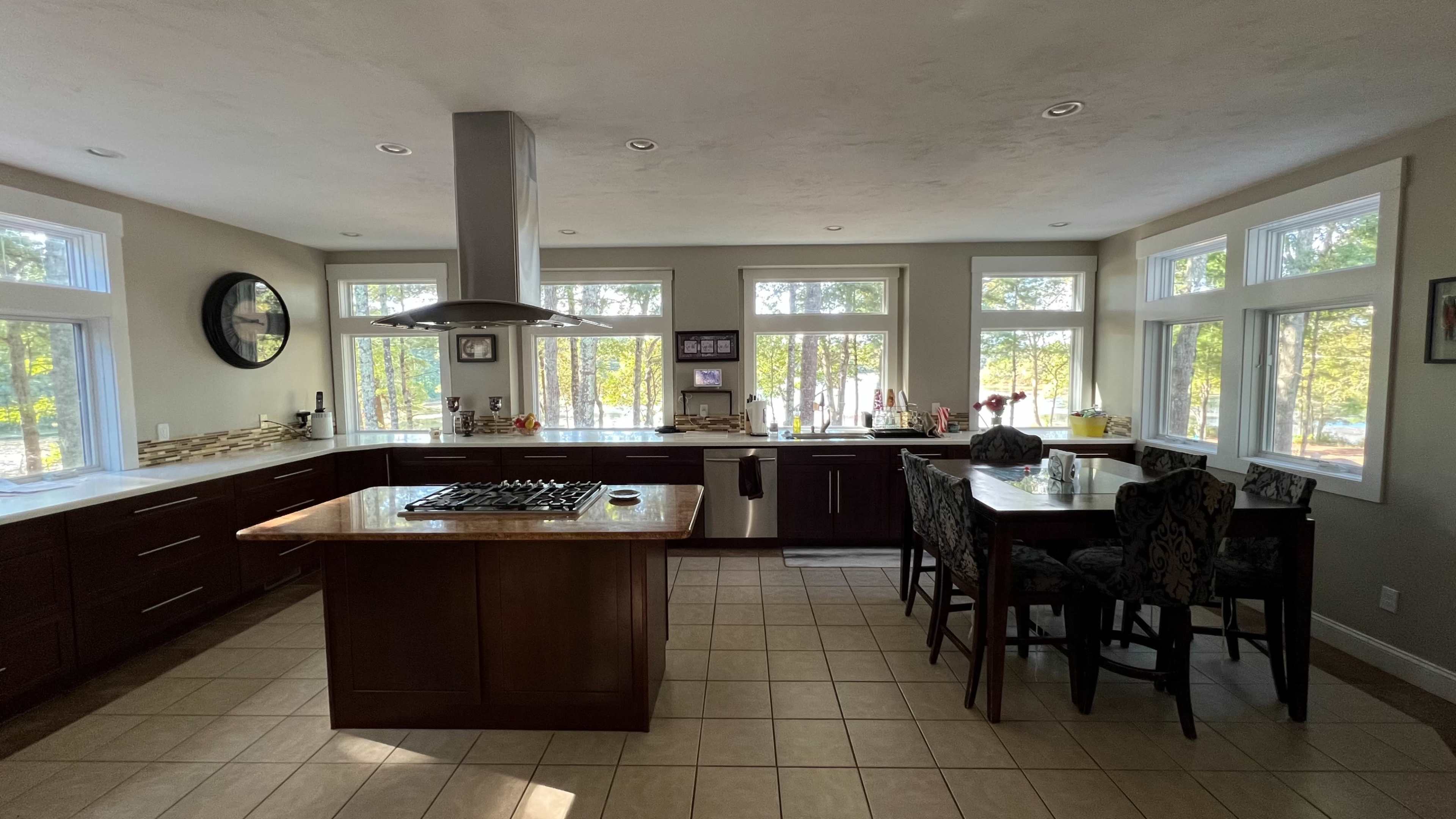 A modern kitchen features dark wood cabinets, an island with a gas stove, and large windows overlooking a wooded area.