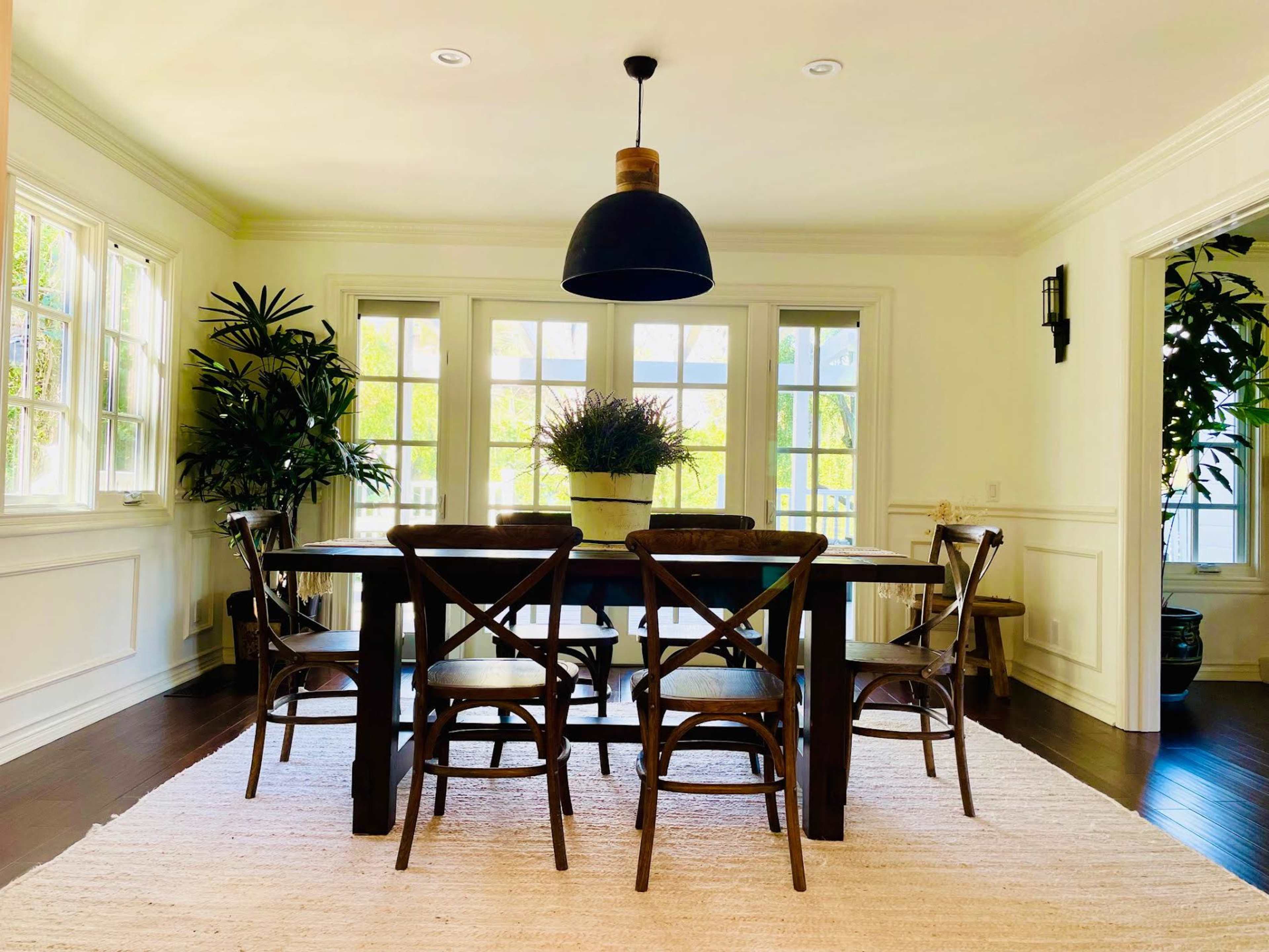 A bright dining area with a large table surrounded by several wooden chairs and a potted plant, illuminated by a black pendant light.
