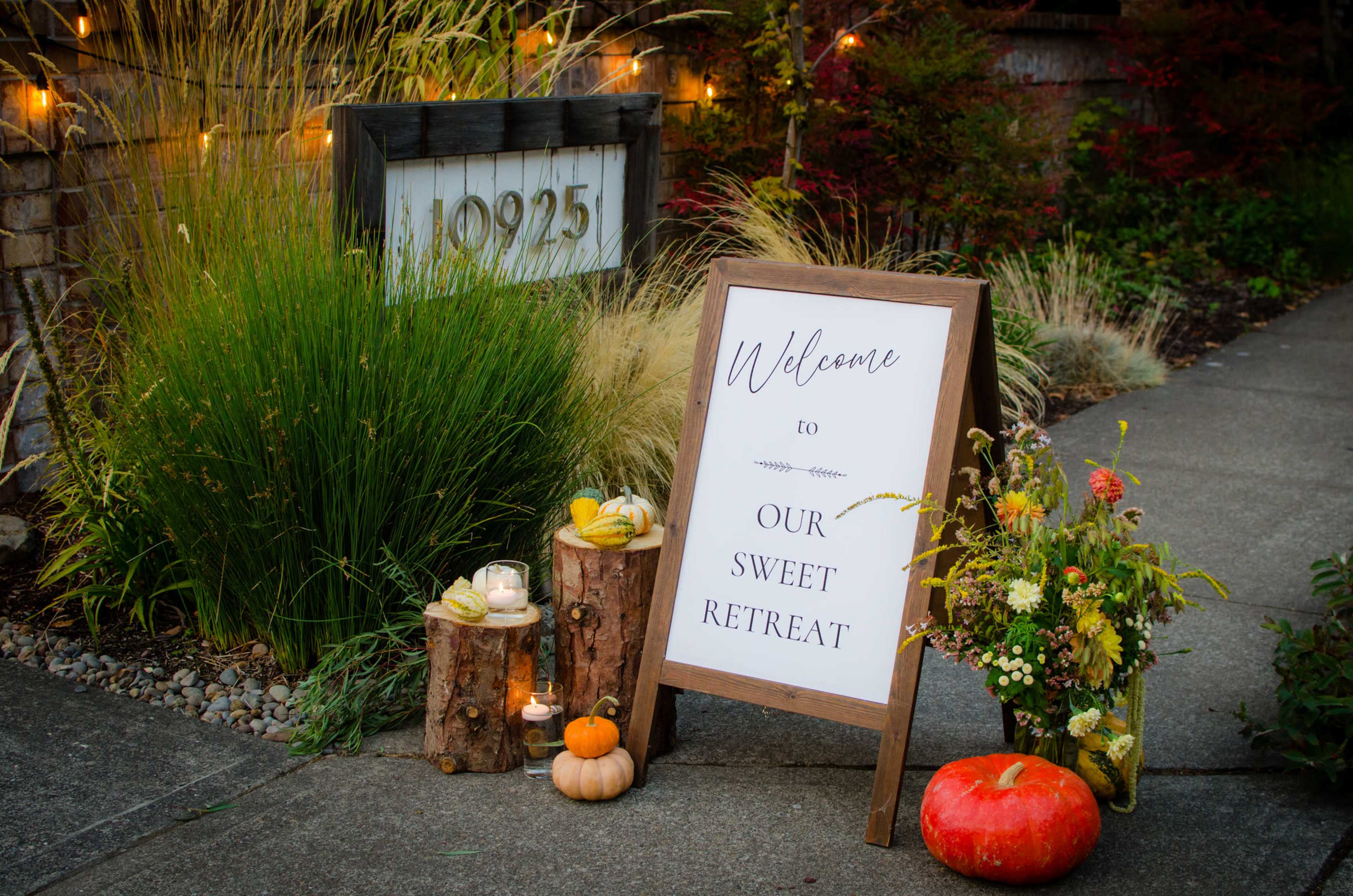A decorative wooden sign welcoming visitors is surrounded by pumpkins, flowers, and logs near a suburban house.