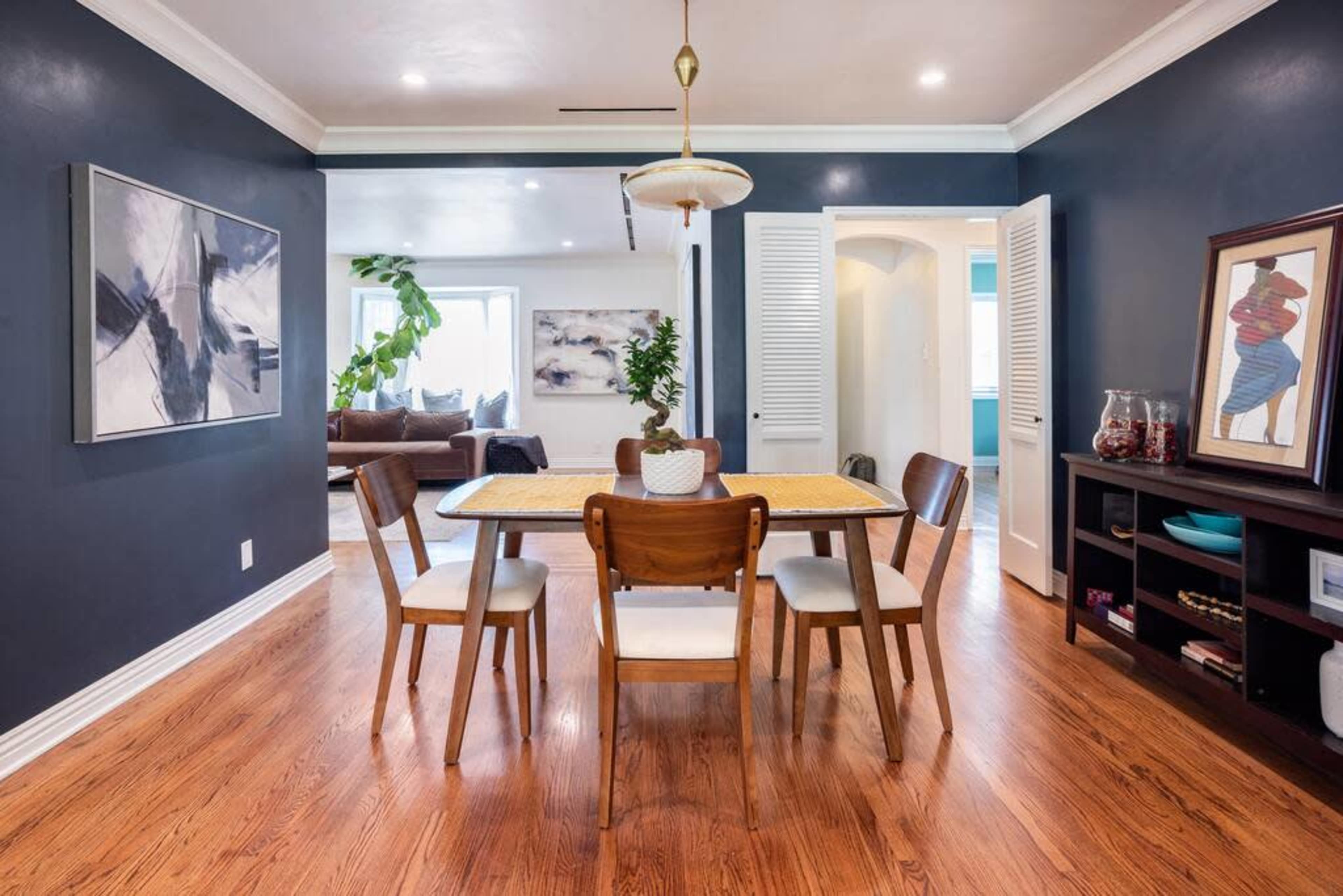 A dining area features a wooden table surrounded by four chairs, with dark blue walls and access to adjoining rooms.
