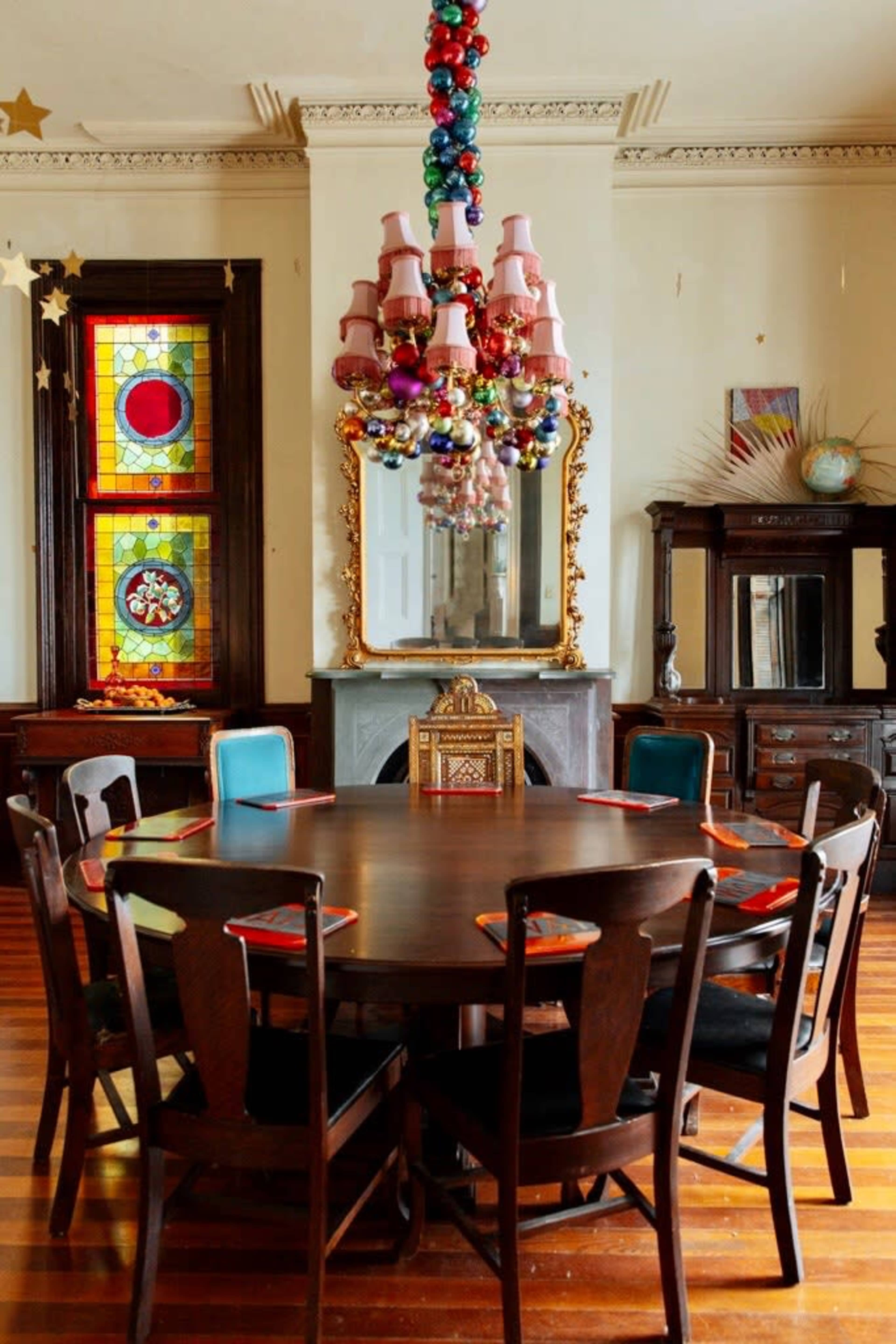 The image features a round dining table surrounded by chairs in a brightly lit room, accented by colorful stained glass windows and a decorative chandelier.