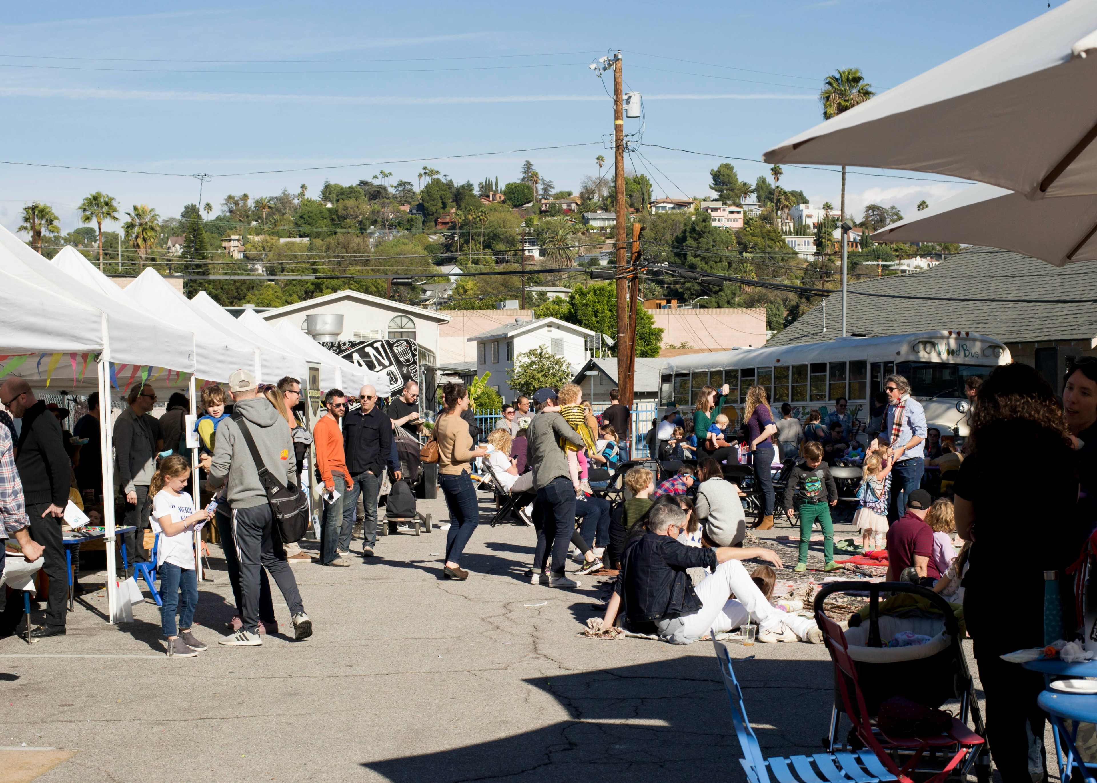 A busy outdoor market scene shows people gathered around booths and sitting on blankets, with tents providing shade and a hillside of houses in the background.