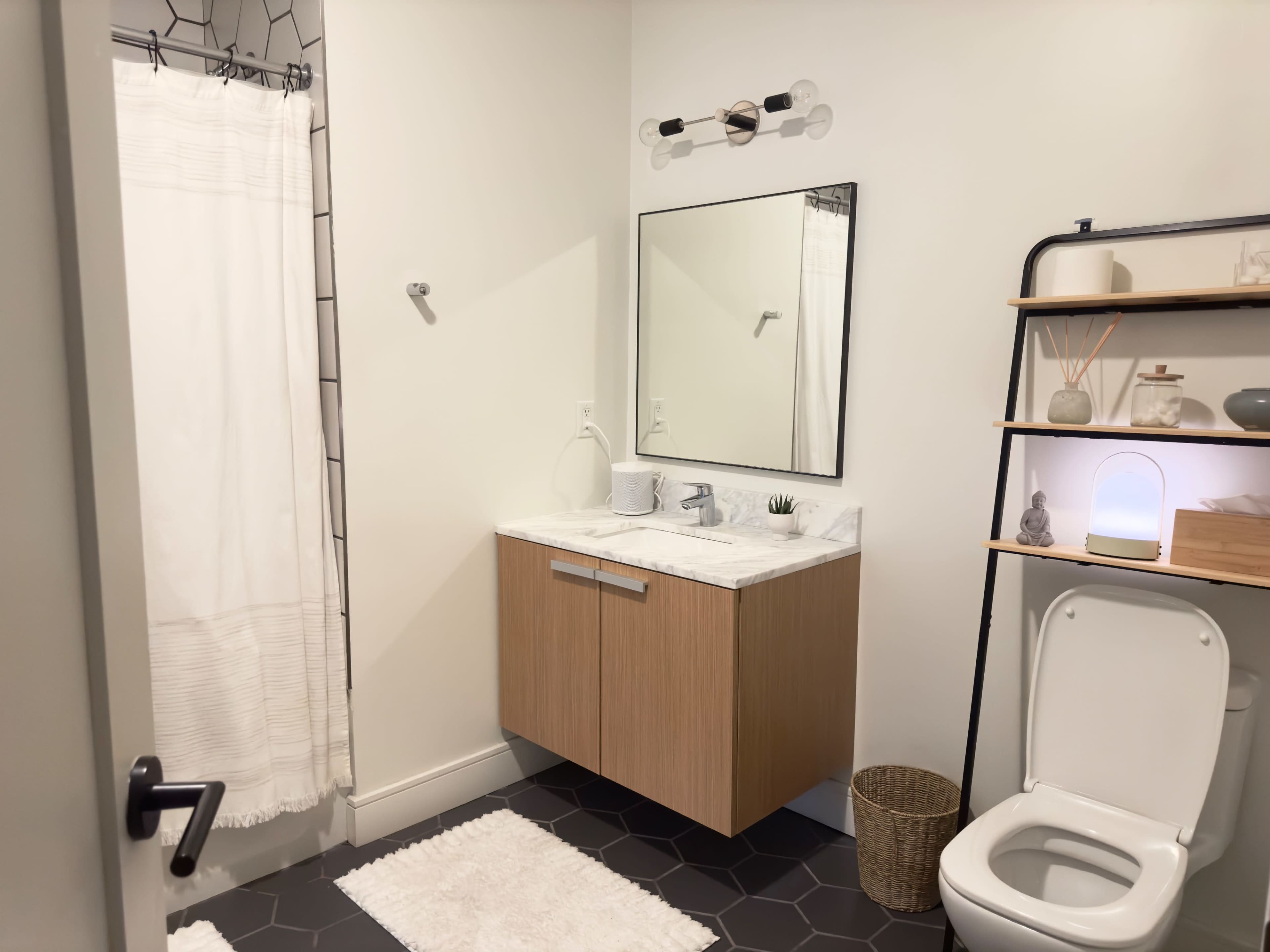 A modern bathroom featuring a wall-mounted mirror above a wooden vanity, a shower with a curtain, and a toilet beside a shelf with decorative items.