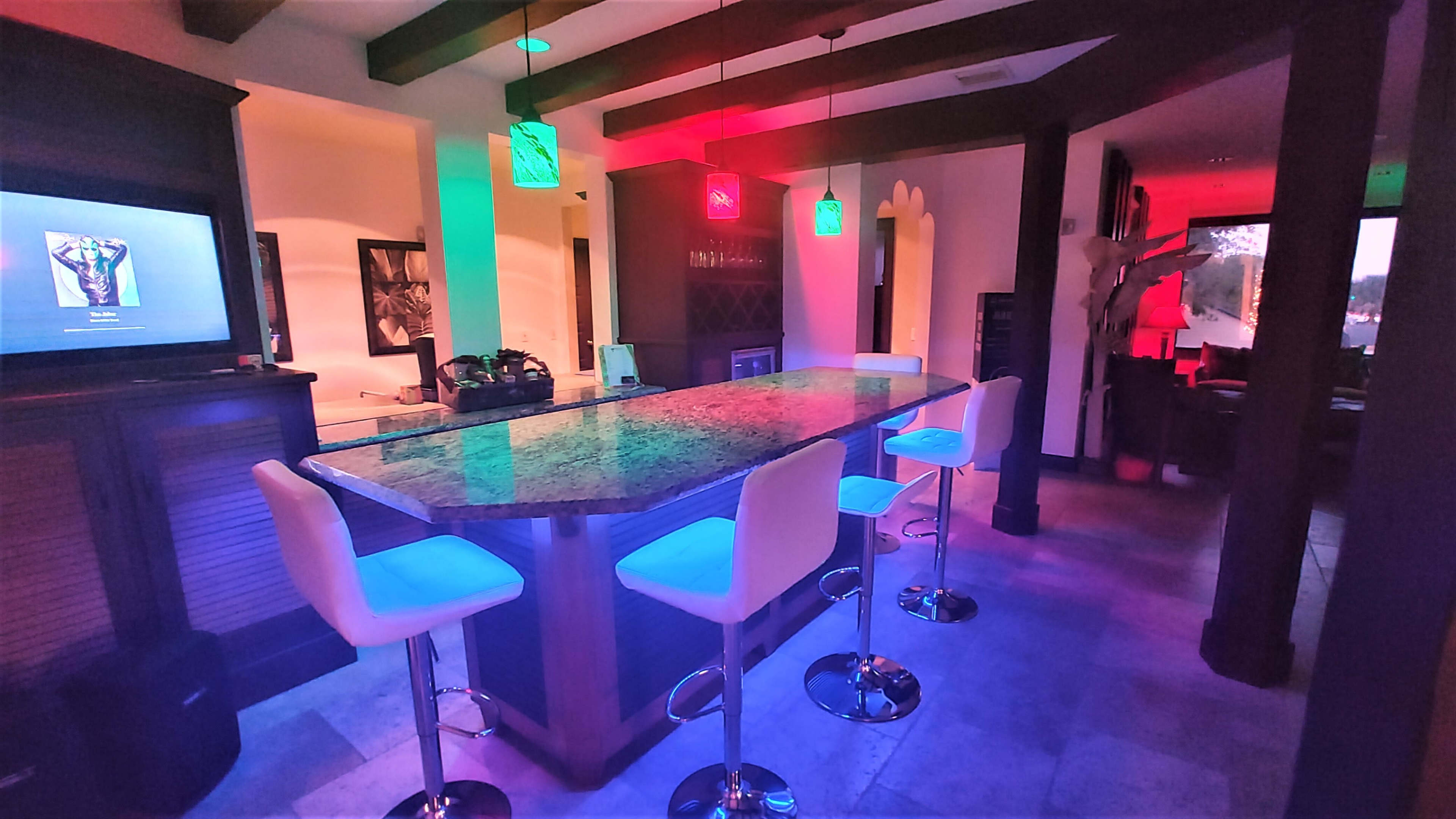 A modern kitchen bar area features a long granite countertop with high stools, accented by colorful pendant lights and a television in the background.