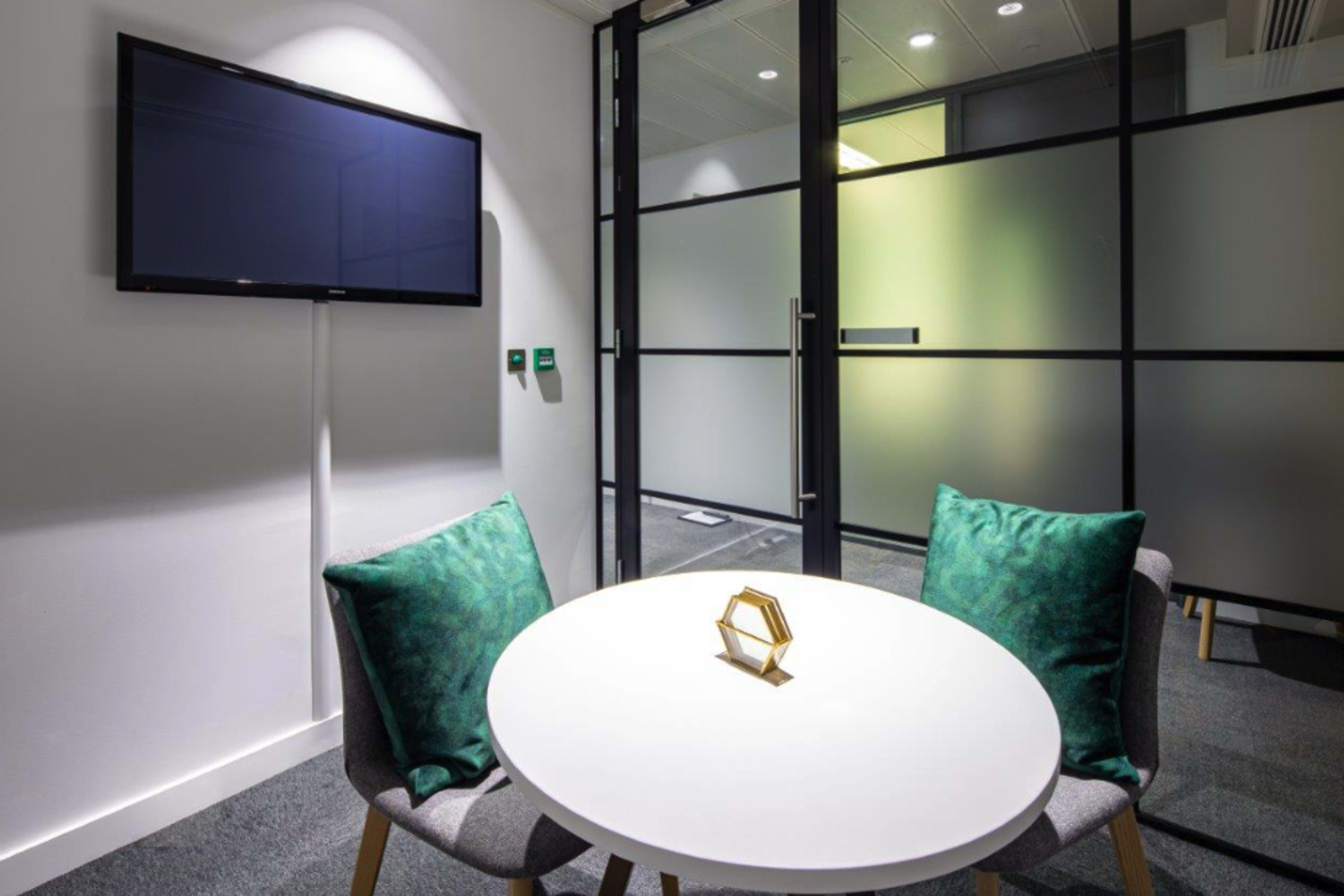 A modern meeting room features a round white table with two green-cushioned chairs and a wall-mounted television.