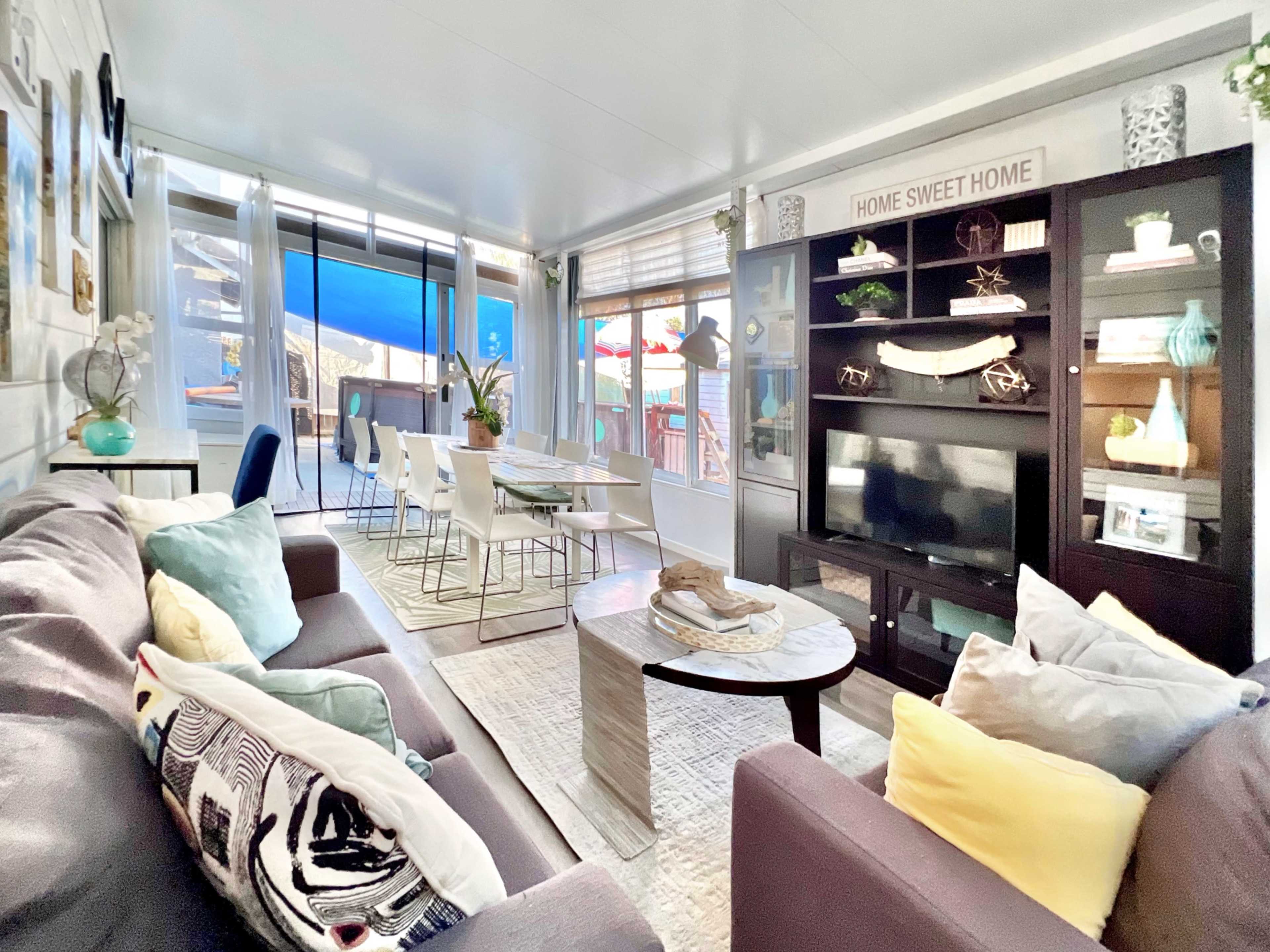 A bright living room features a comfortable couch, a central coffee table, a television stand with decorative items, and a dining area adjacent to large windows.