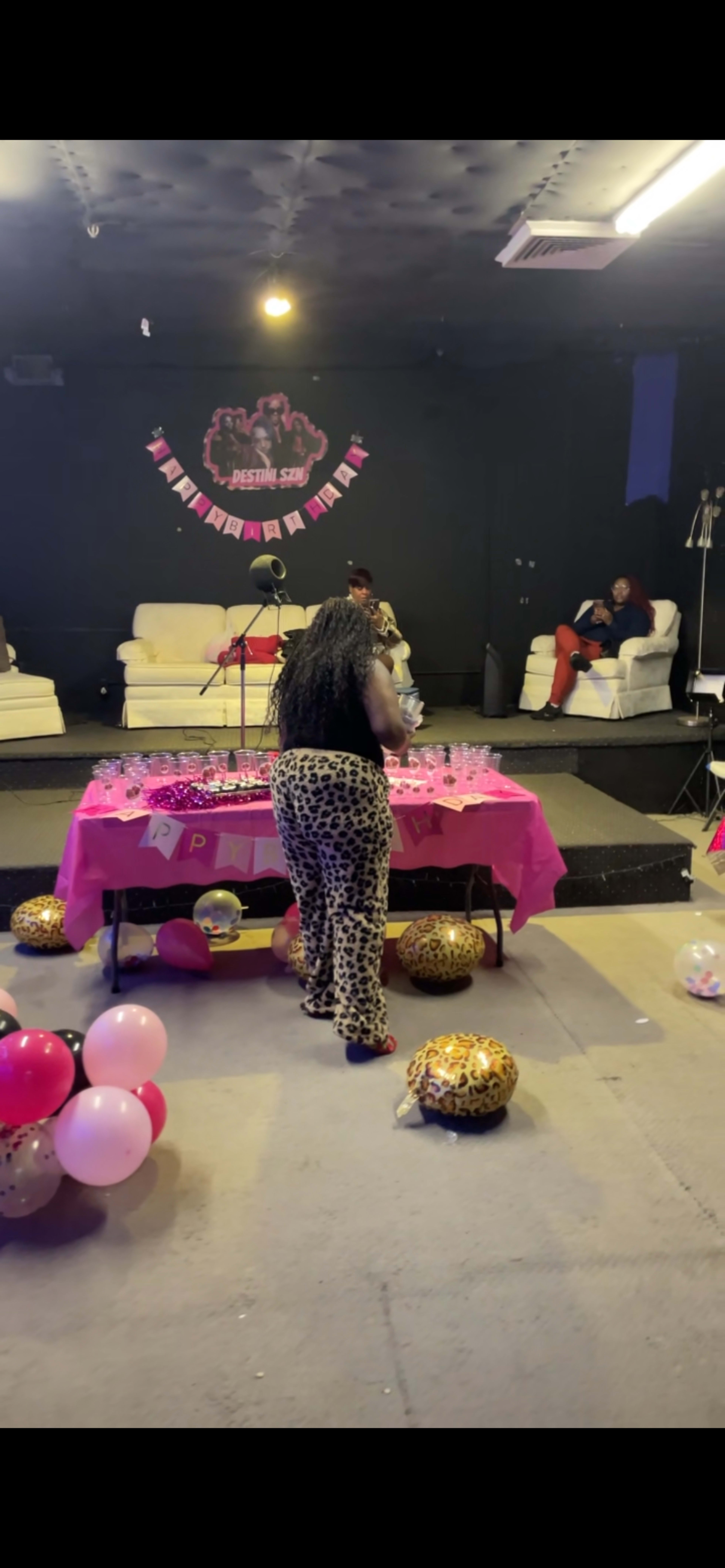 A woman in leopard print pants prepares a table decorated with pink items and balloons in a dimly lit party venue.