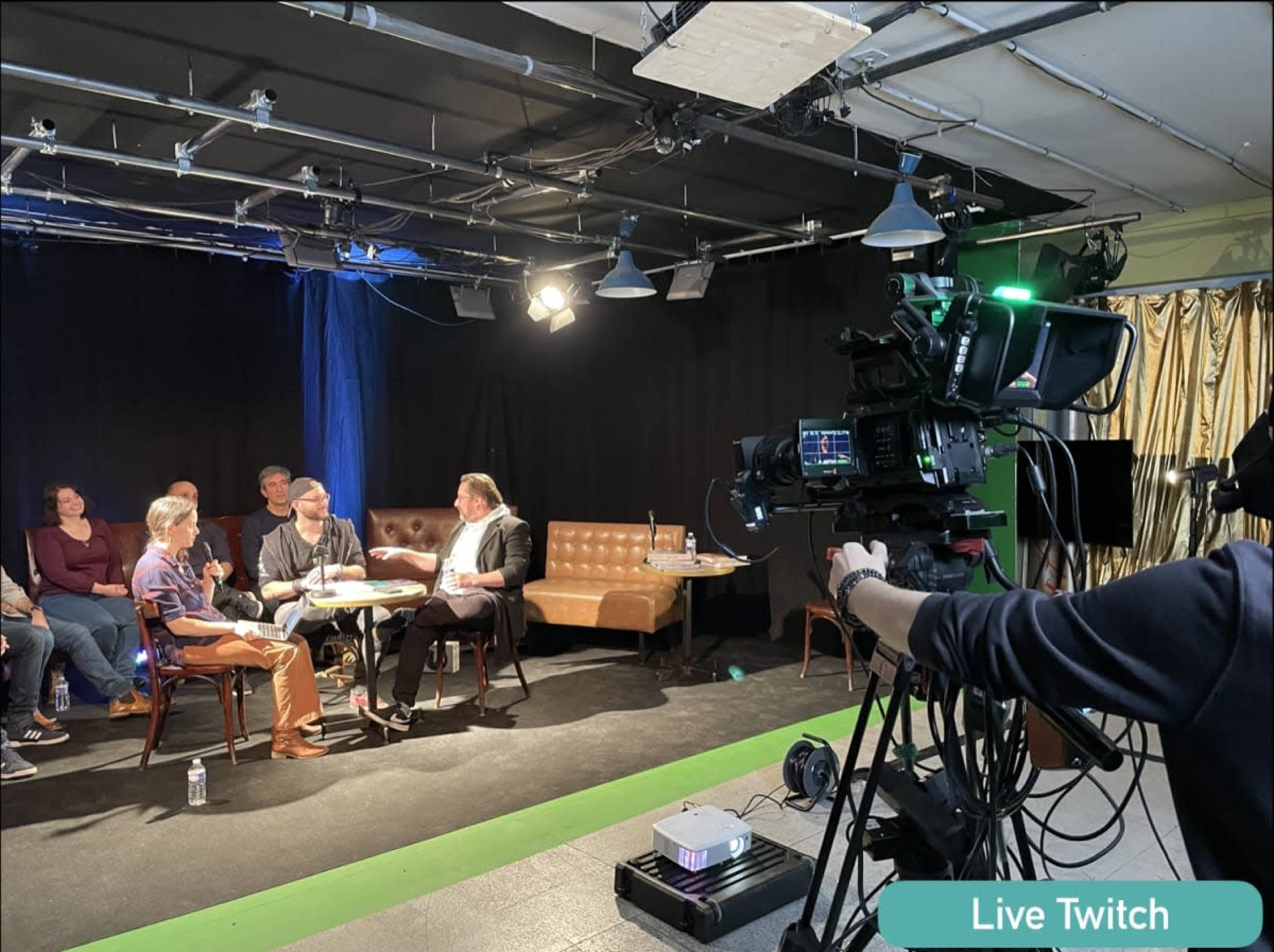 A video production setup captures a discussion on a stage with chairs and a camera focused on participants seated at a table.