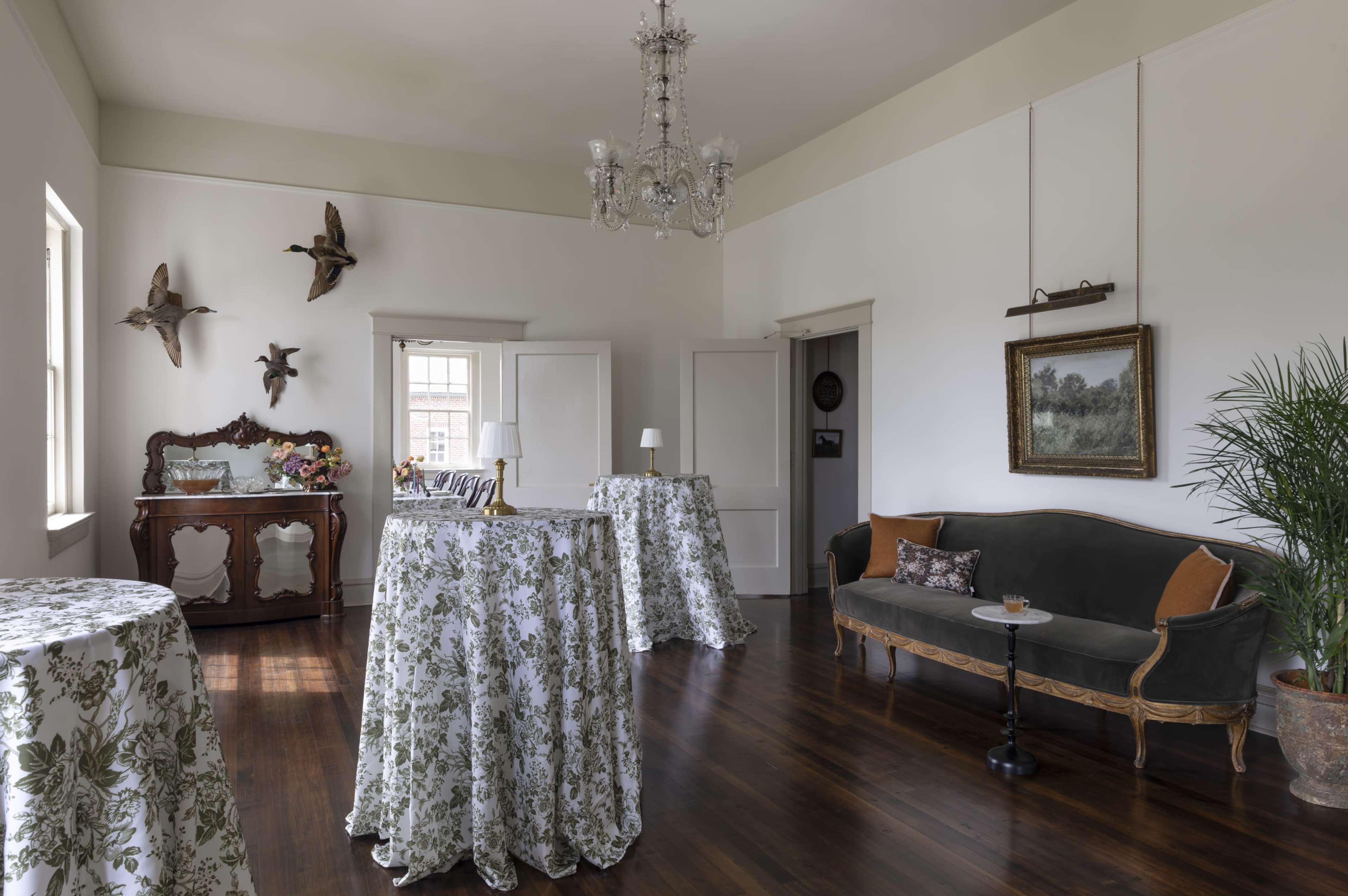 The interior of a room features wooden flooring, a vintage-style sofa, and several small tables draped with floral-patterned cloth, along with a chandelier and decorative wall art.