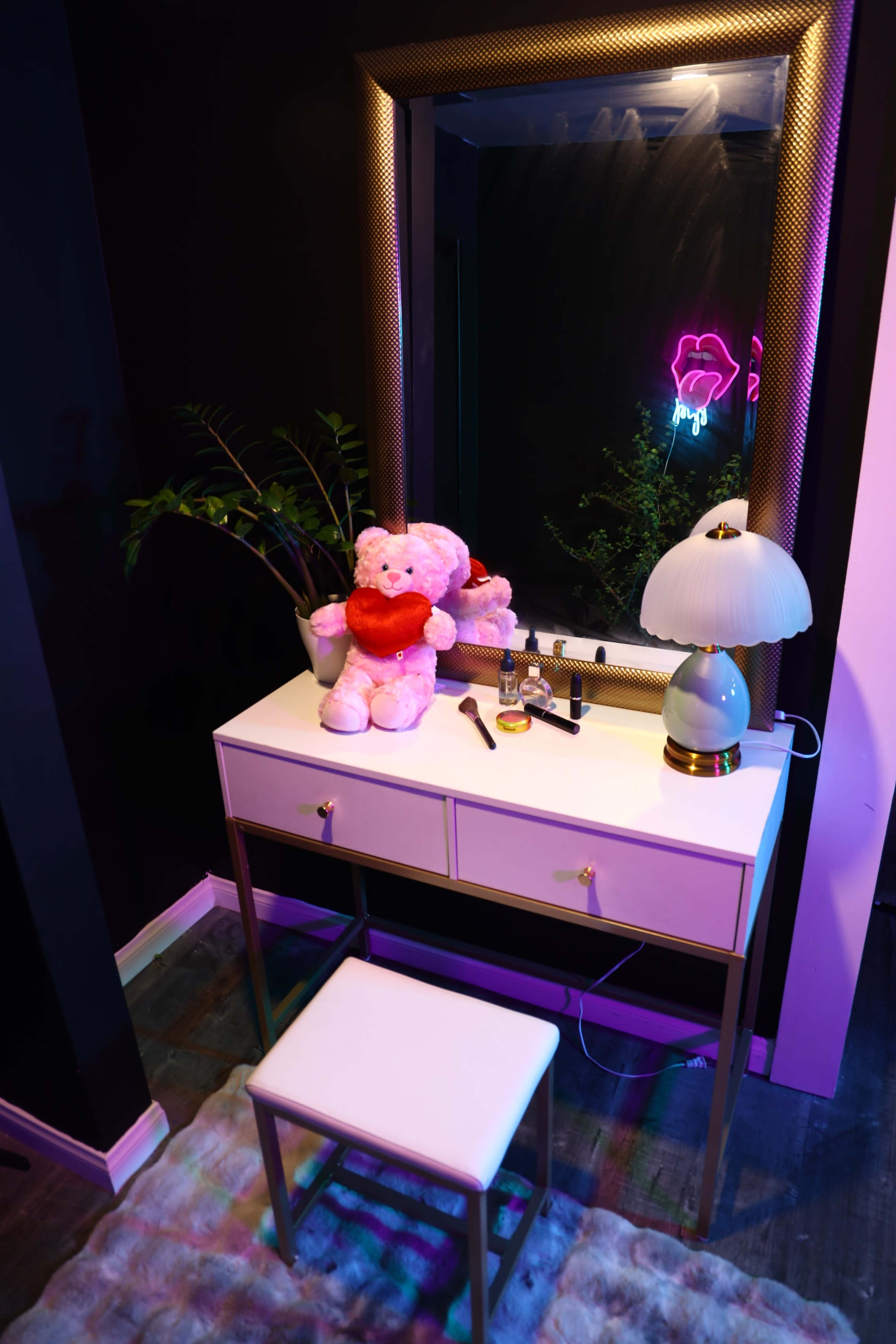 A vanity table with a mirror, a stuffed bear, a lamp, and various cosmetics is set against a dark wall with a neon light.