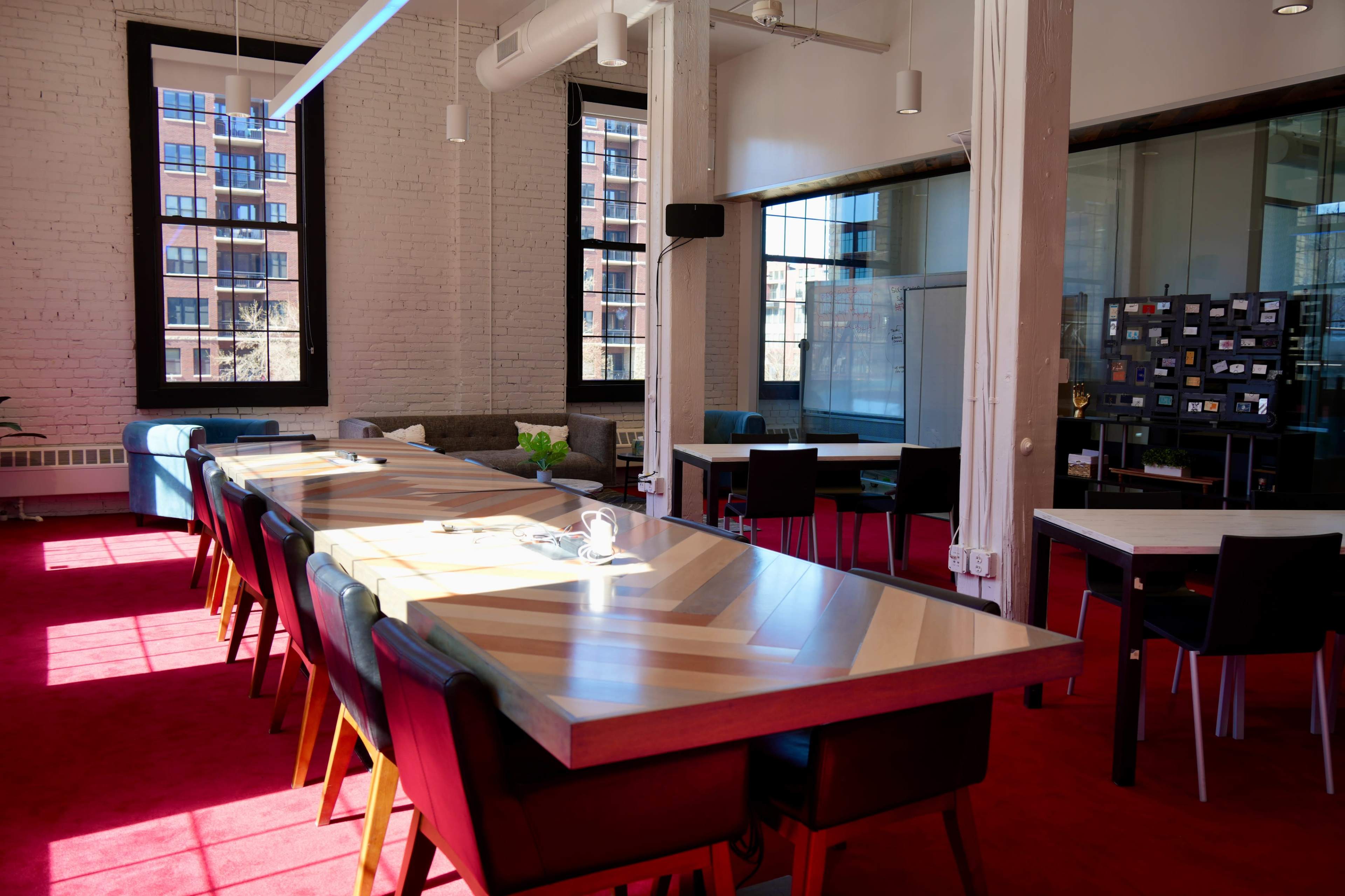 The image shows a bright, modern workspace featuring a long, multi-colored table with various chairs, surrounded by large windows allowing natural light to illuminate the red carpeted floor.