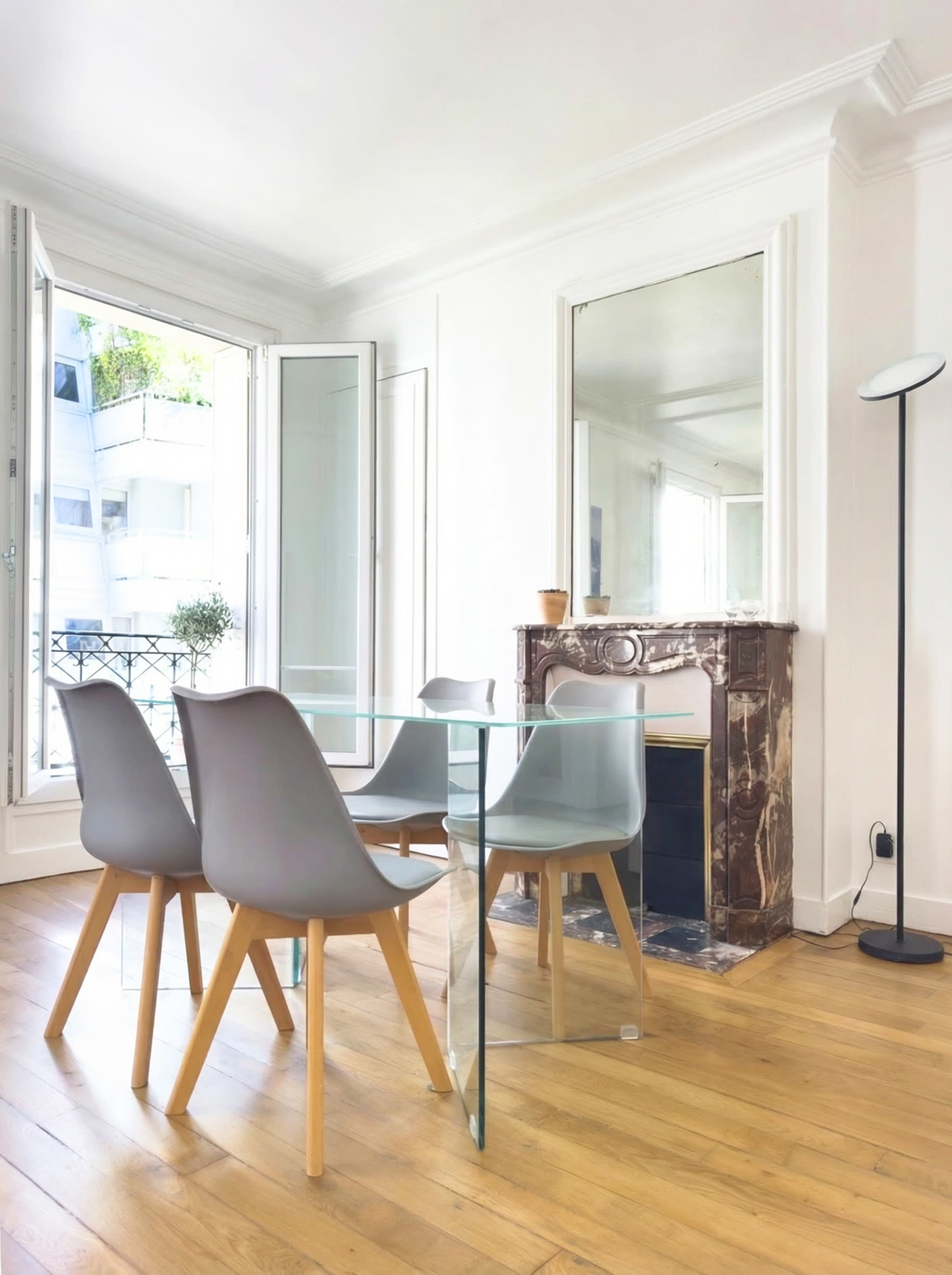 The image shows a modern dining area with a glass table and four gray chairs, complemented by a fireplace and large windows allowing natural light to fill the space.