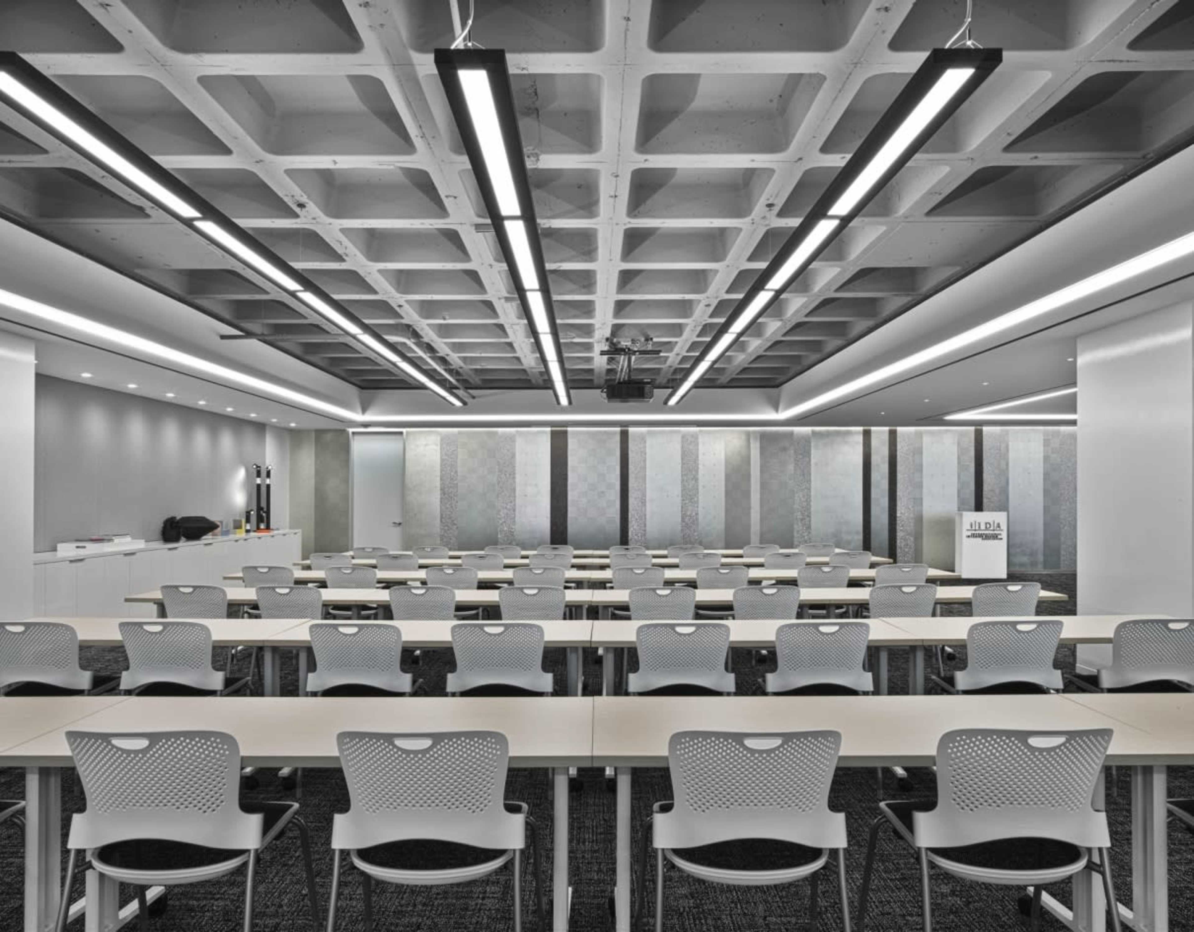 The image shows a modern conference room set up with rows of empty desks and chairs under a structured ceiling with fluorescent lighting.