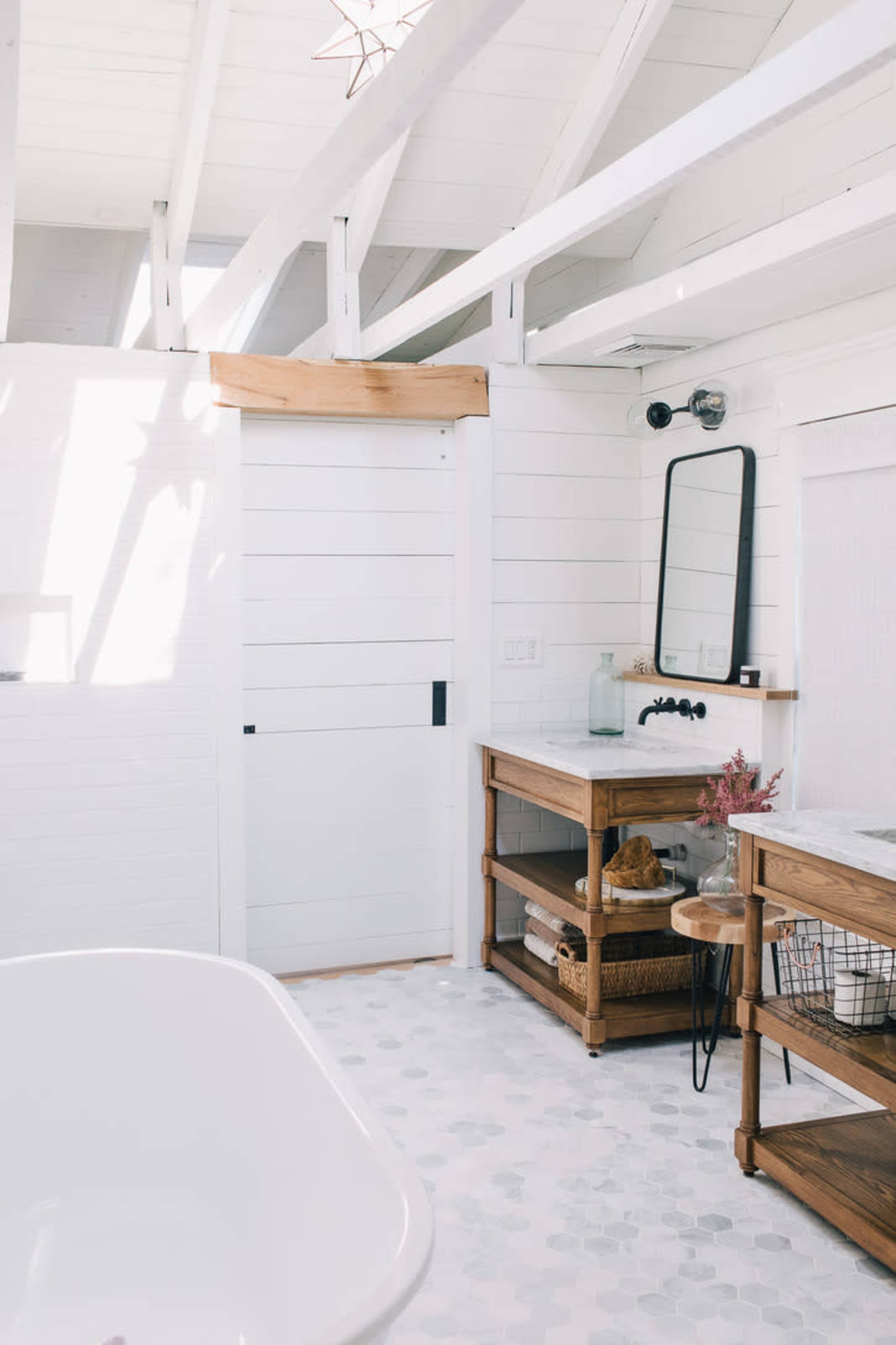 The image shows a modern bathroom with wooden accents, a freestanding bathtub, and a large mirror above a wooden vanity near a white sliding door.