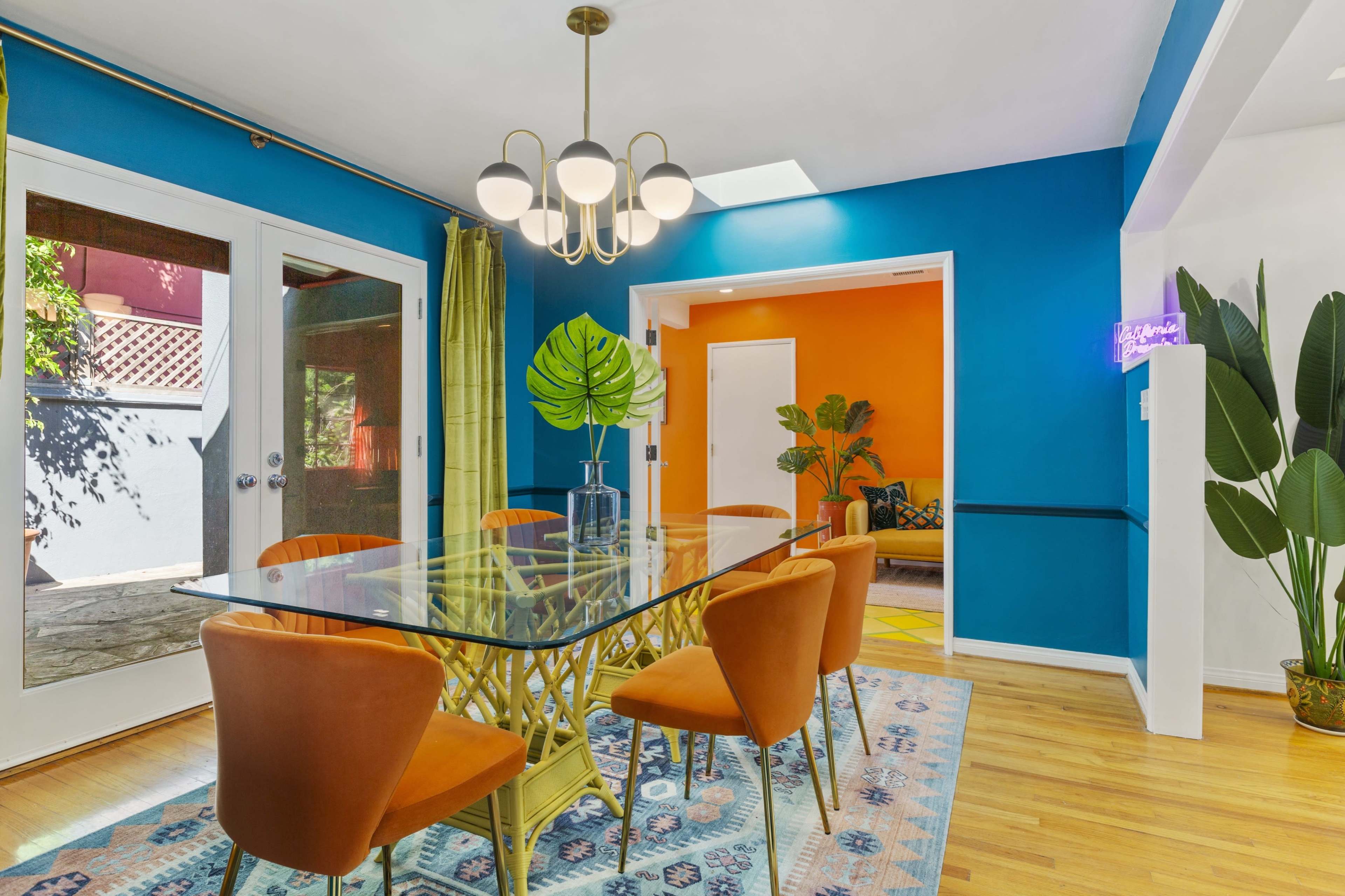 A dining room features a glass table surrounded by orange chairs, with vibrant blue and orange walls and large plants in the corners.