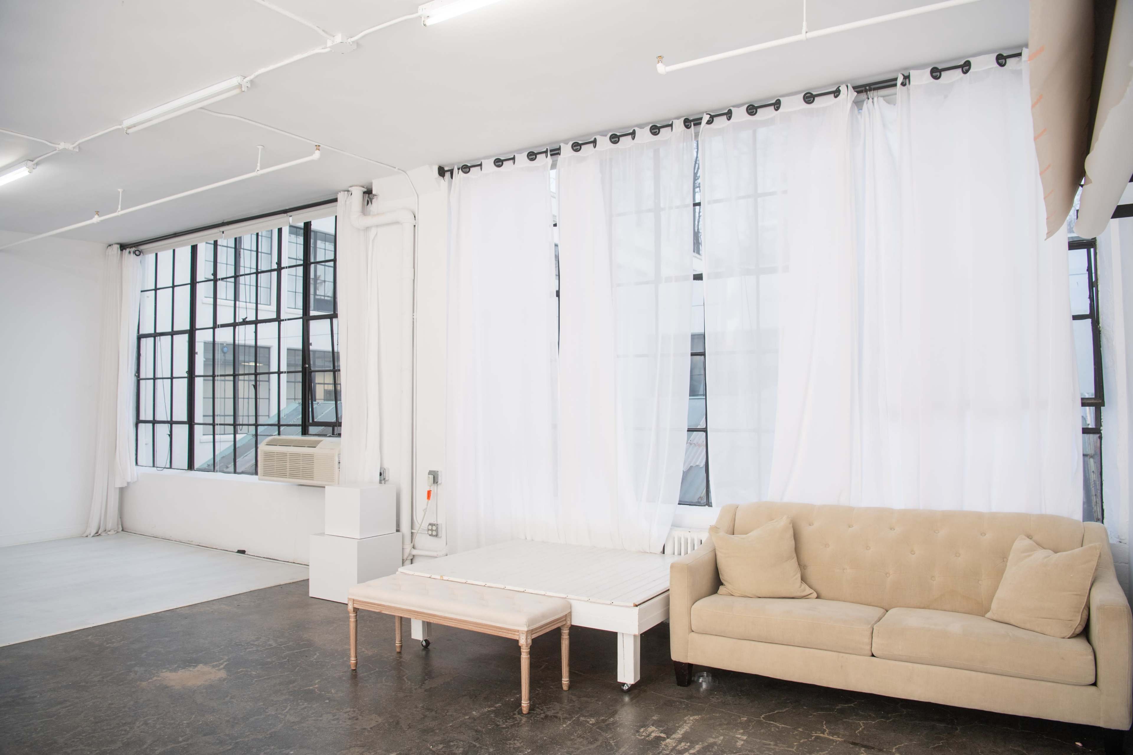 An interior space with large windows, white sheer curtains, a beige couch, and a small wooden table on a dark floor.