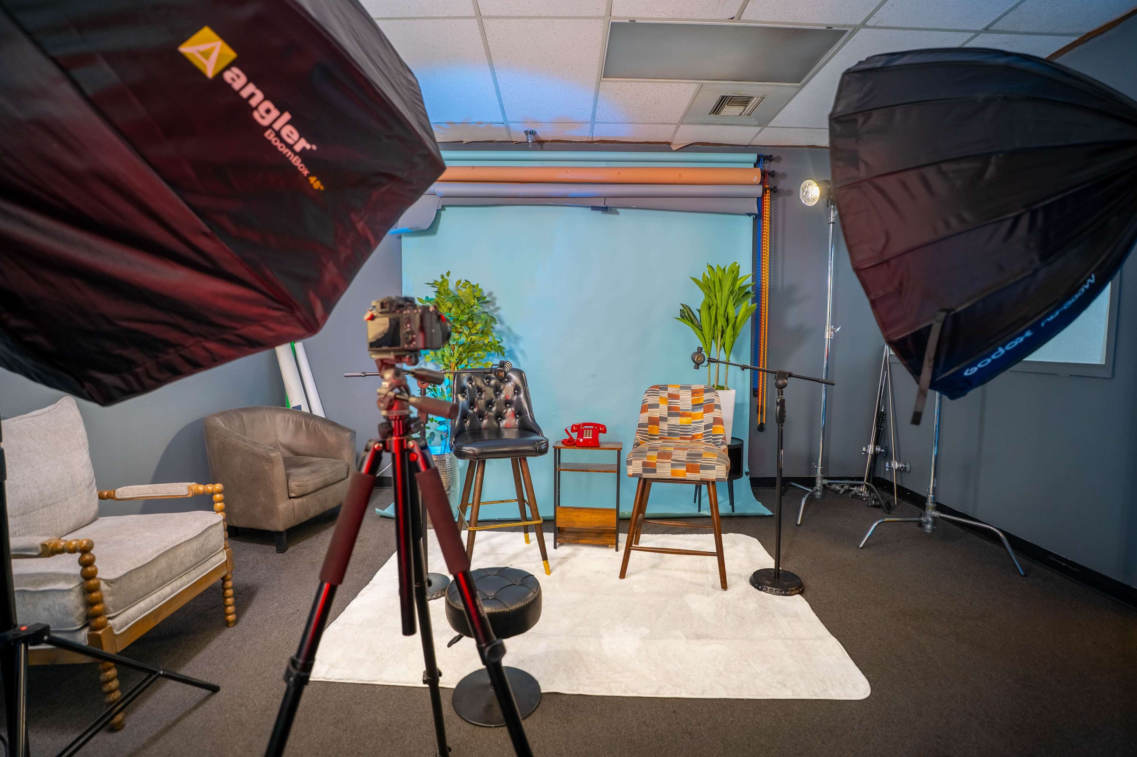 A photography studio is arranged with two chairs, a camera on a tripod, and large softbox lights, all set against a blue backdrop.