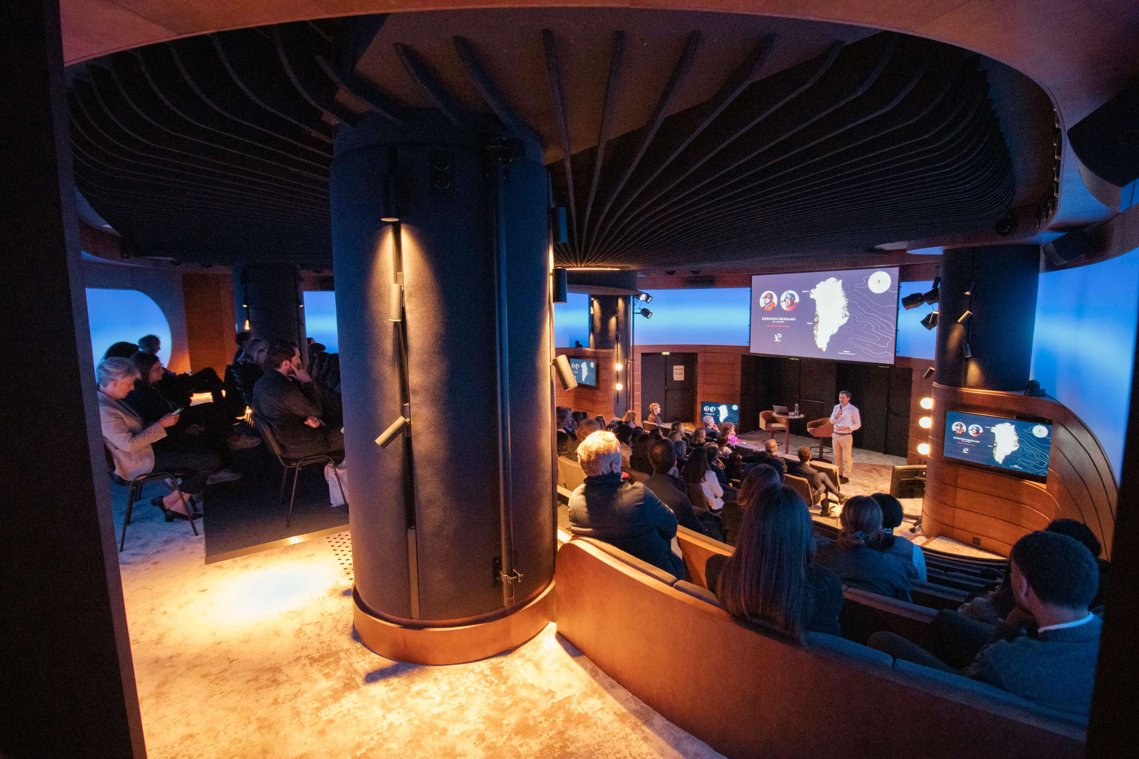 A presentation is taking place in a modern auditorium with a curved design, where an audience is seated and facing a large screen displaying information.