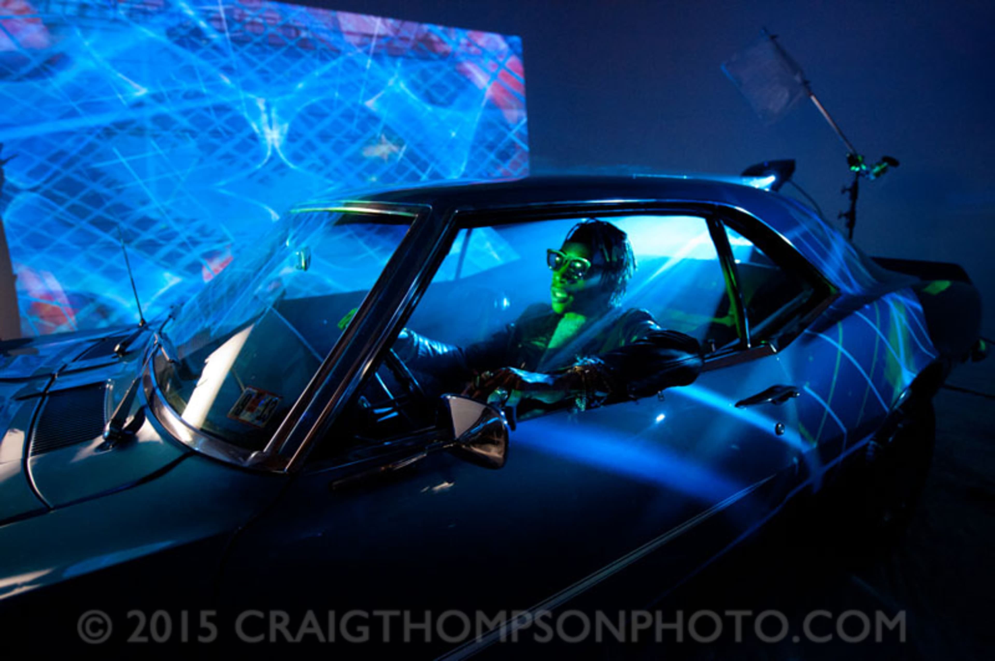 A person sits in the driver's seat of a car illuminated by colorful light projections in a dimly lit environment.