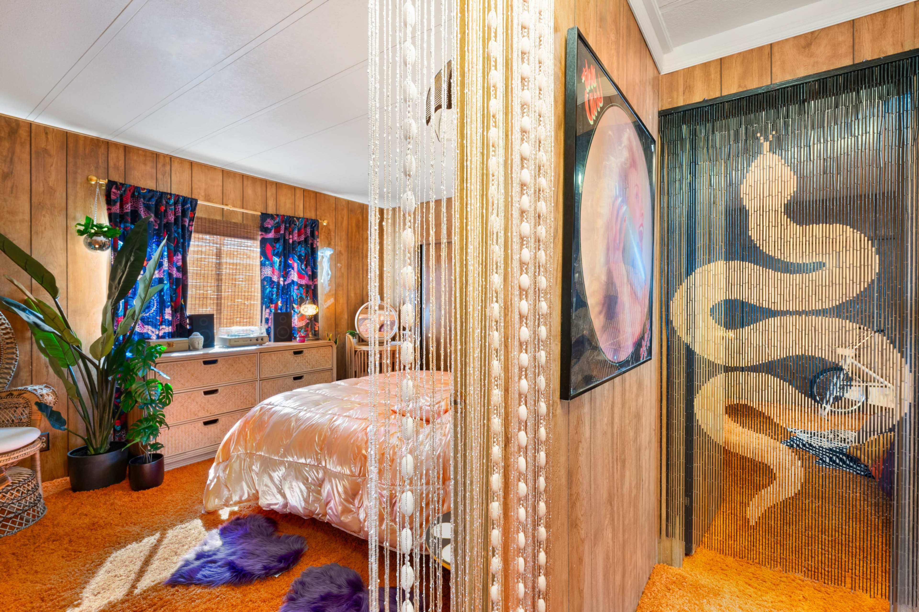 The image shows a brightly decorated bedroom featuring a bed with metallic bedding, wood-paneled walls, and decorative curtains, with a beaded curtain leading to another space.