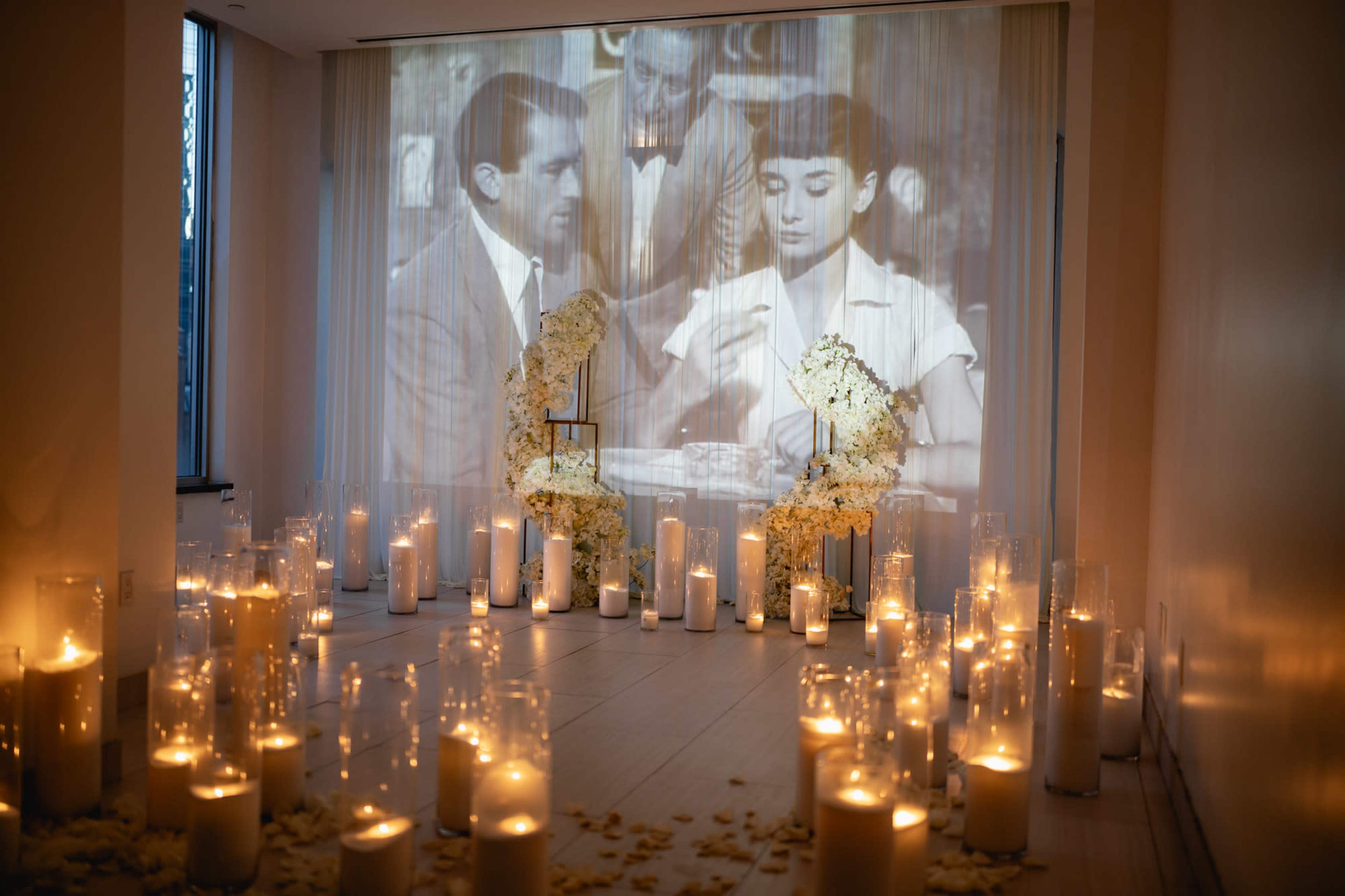 A dimly lit room features a vintage movie projection on a wall, surrounded by white candles and floral arrangements on the floor.