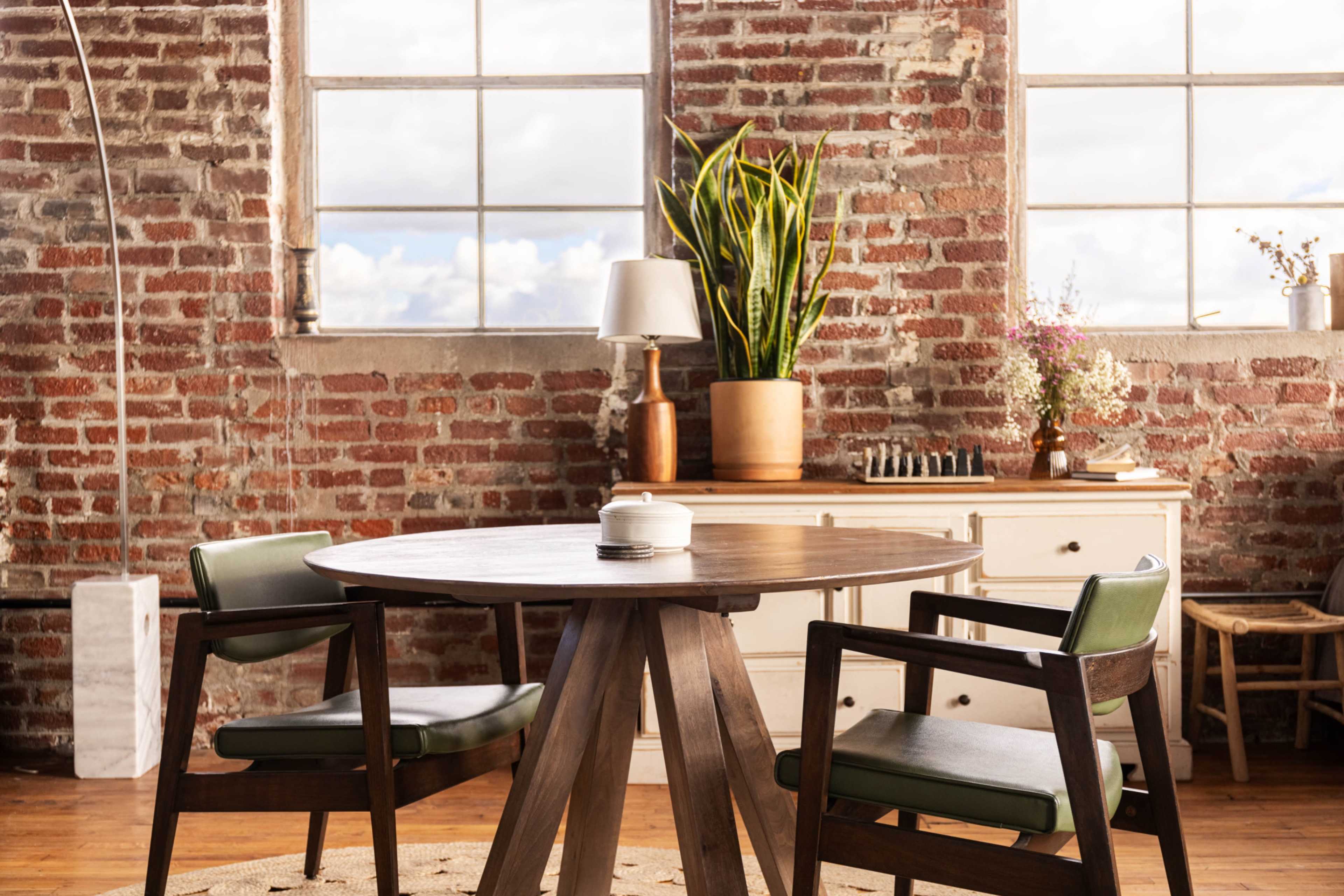 A dining table with two chairs, a lamp, and potted plants in a room with exposed brick walls and large windows.