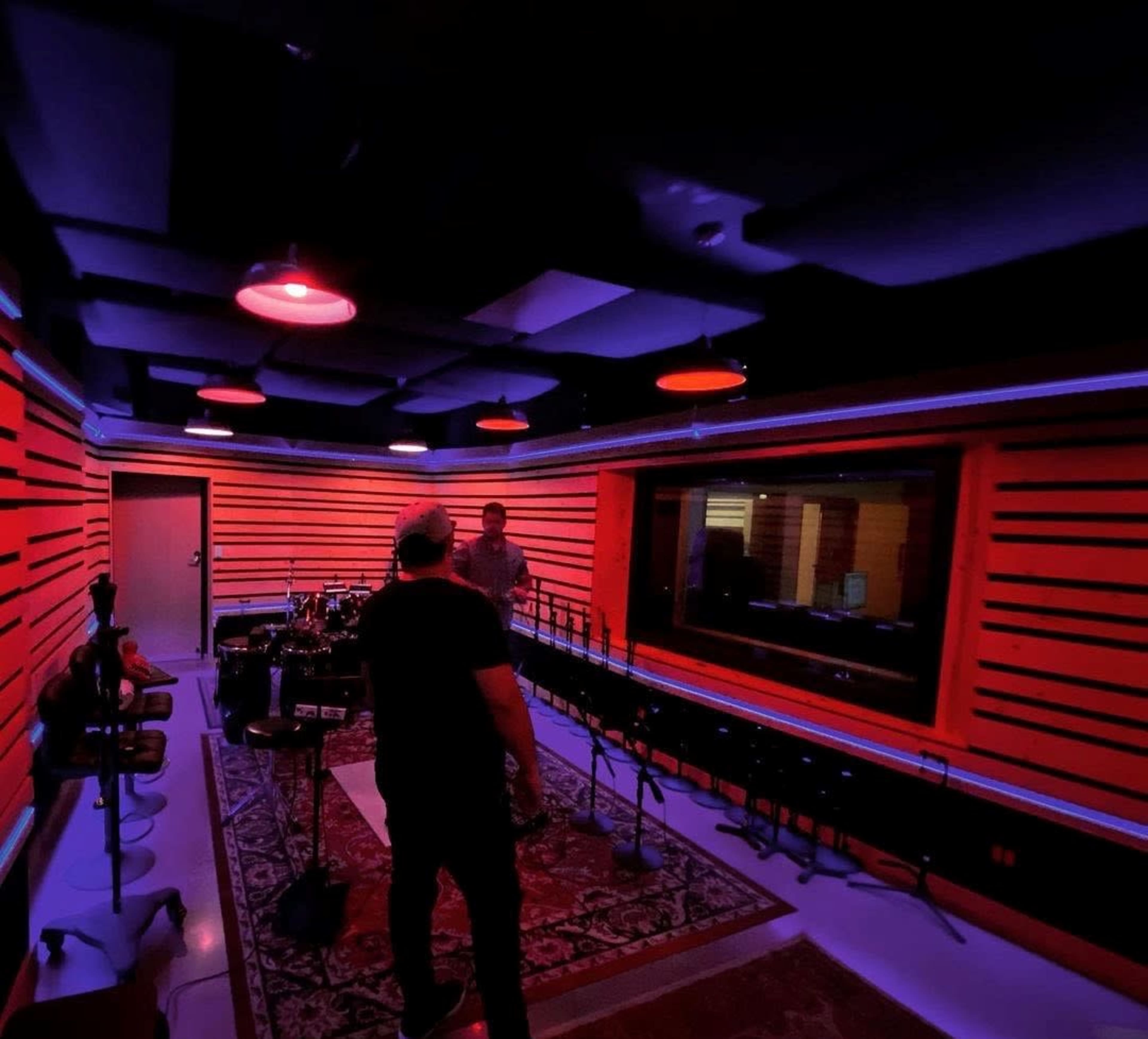 A dimly lit recording studio features red accent lighting, wooden wall panels, and multiple microphones set up in front of a large glass window.
