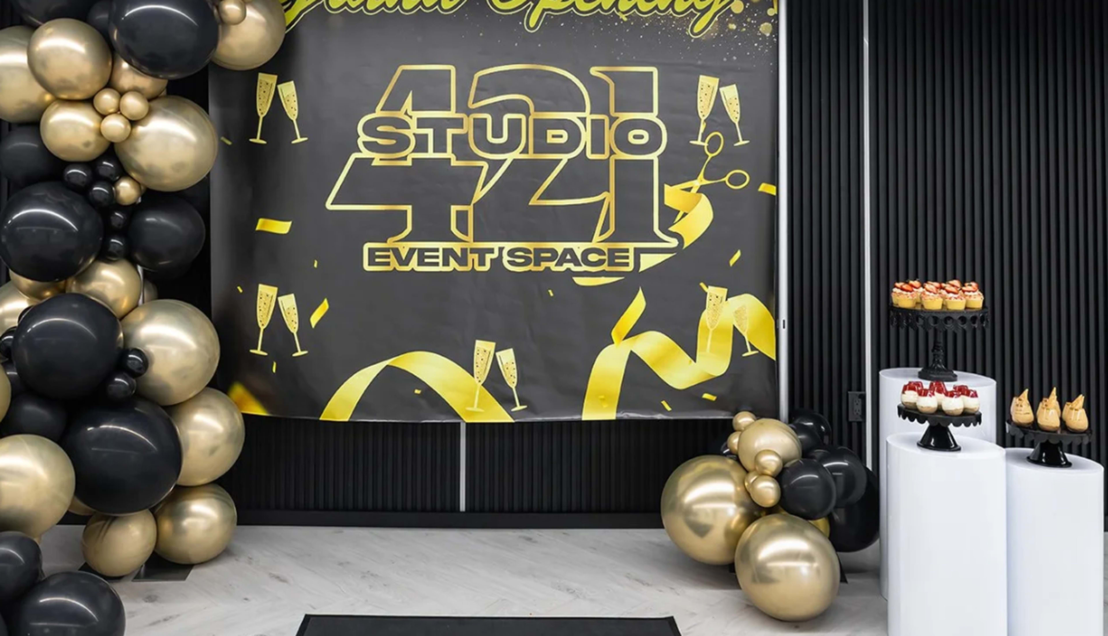The image shows a grand opening setup for "Studio 421 Event Space," featuring a large black and gold banner, a decorative arrangement of black and gold balloons, and a display of desserts on stands.