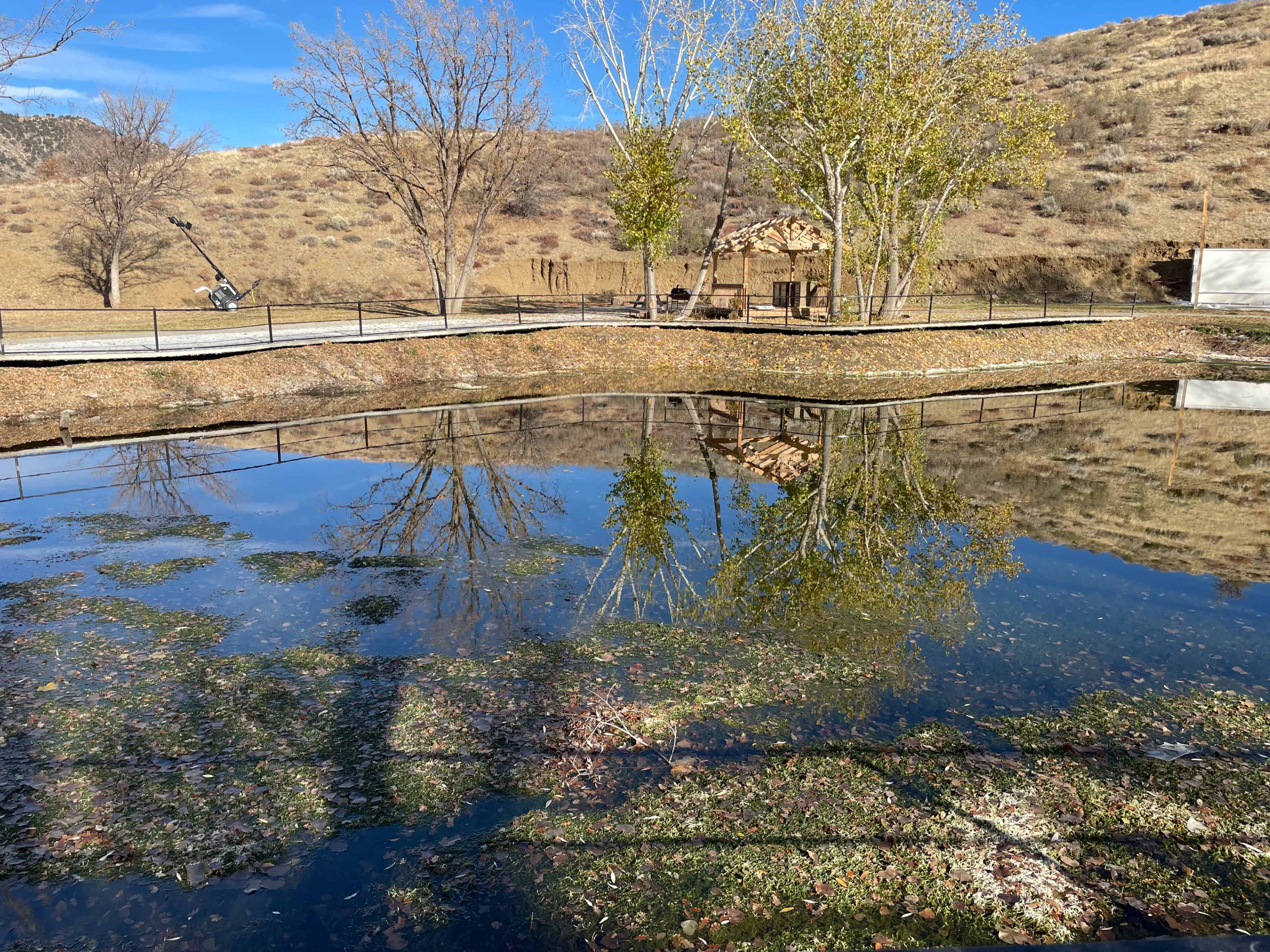 A calm pond reflects trees and a small structure, surrounded by dry hills and a clear sky.