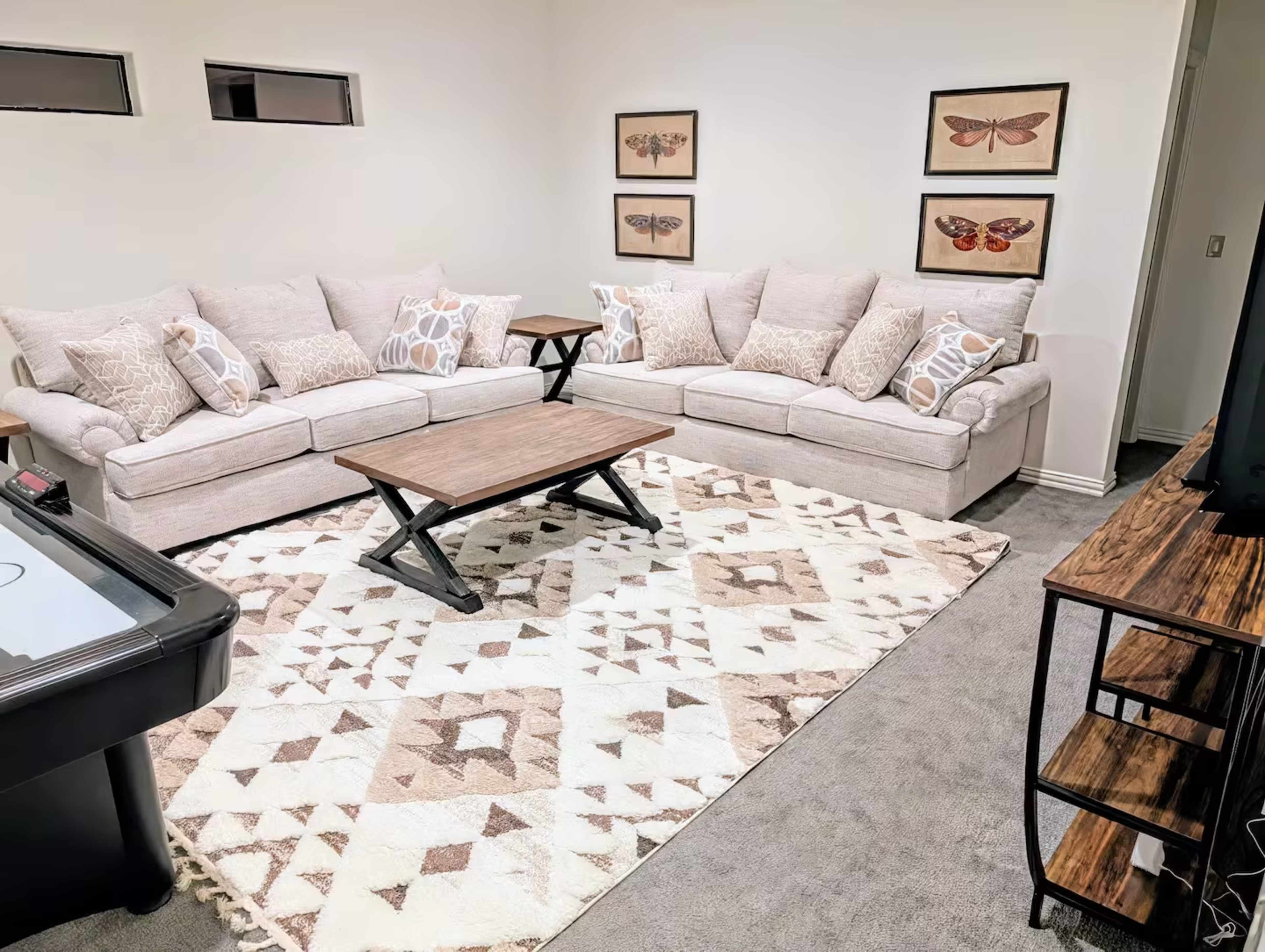 The image shows a cozy living room with a large sectional sofa, a wooden coffee table, and decorative butterfly art on the walls, along with a rug on a gray carpeted floor.